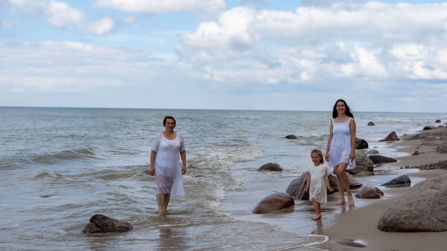 Three generations of women in white dresses wade along a rocky shoreline under cloudy skies.