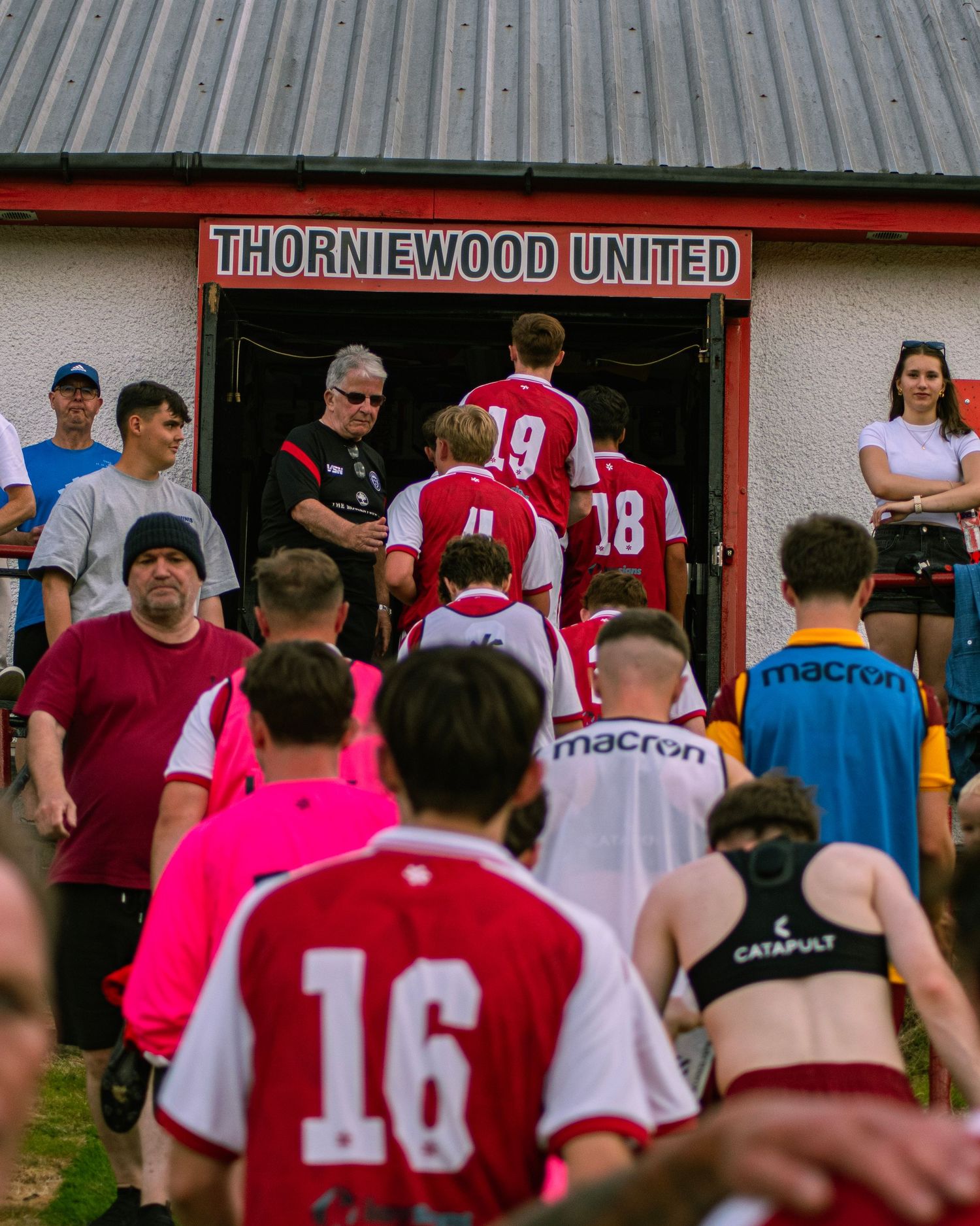 Players in red and white Thorniewood United jerseys entering clubhouse during a football match day.
