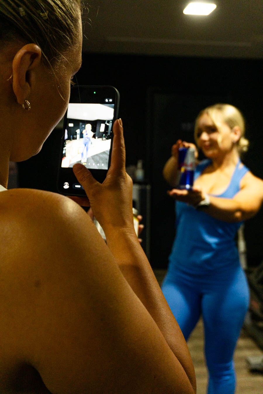 Woman in blue athletic wear holds a water bottle while being photographed on a smartphone in a gym.