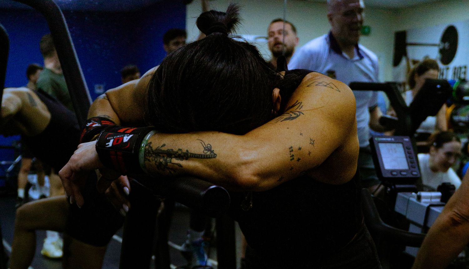 Tattooed athlete resting head on arms during intense gym workout session surrounded by fitness equipment.