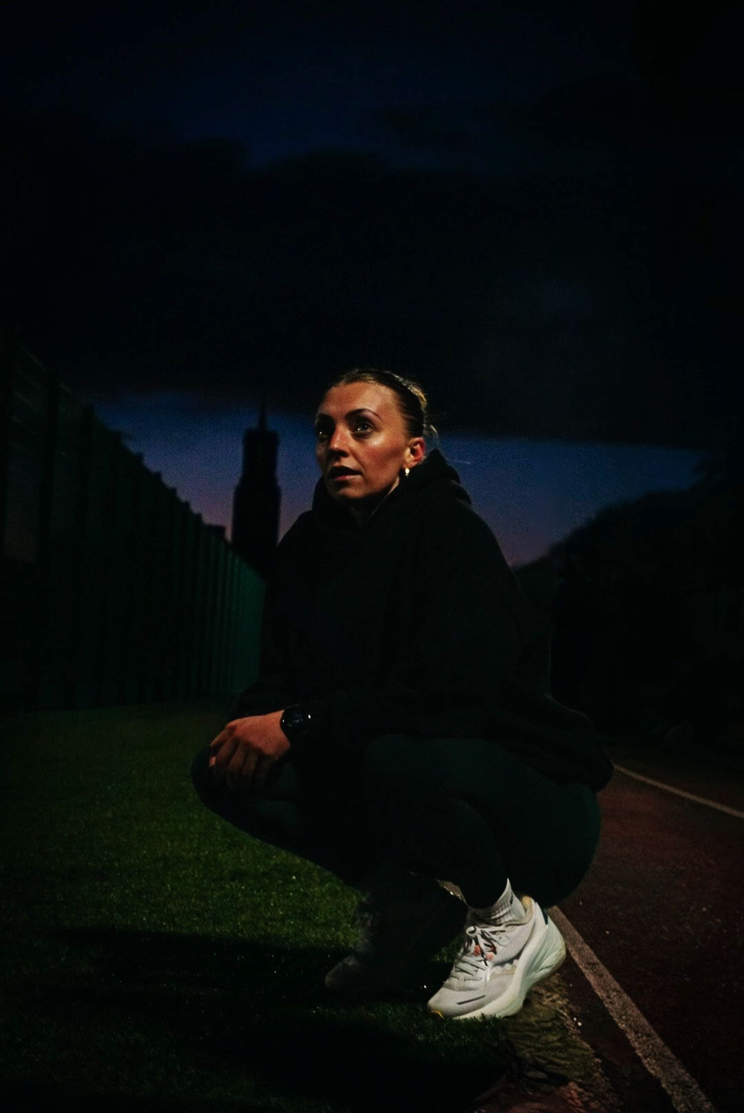 Female athlete crouching outdoors at dusk wearing a black hoodie and white sneakers.