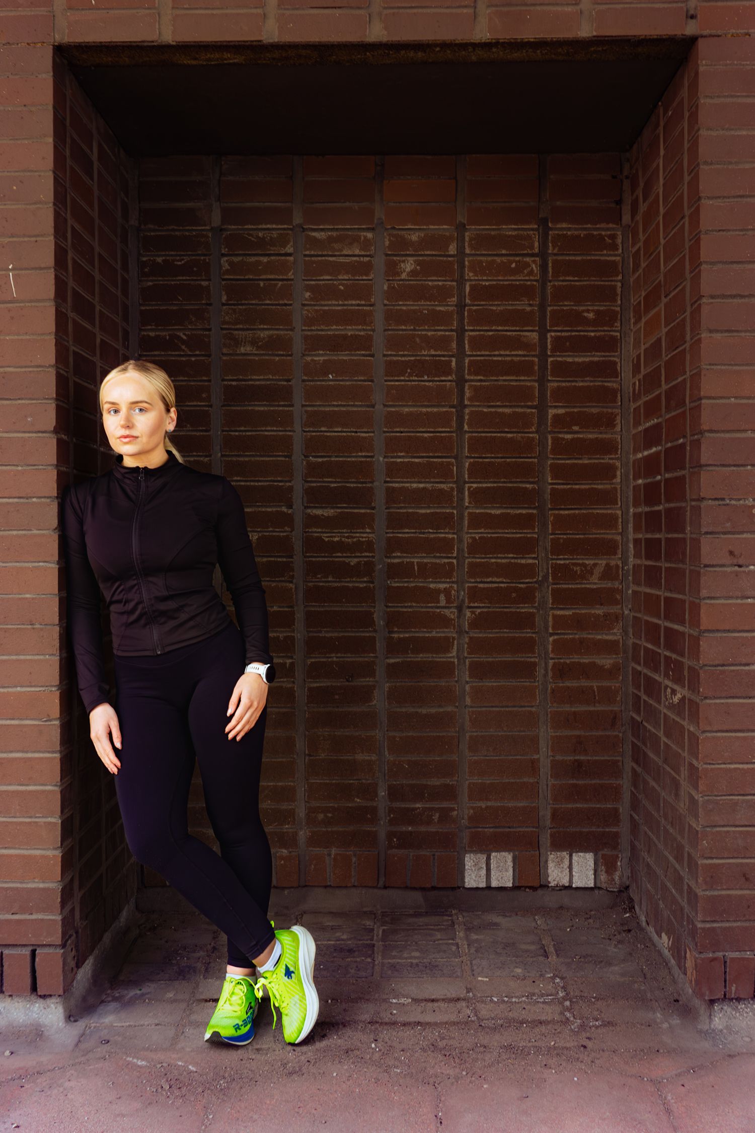Female athlete in black sportswear leaning against brown brick wall wearing neon green sneakers.