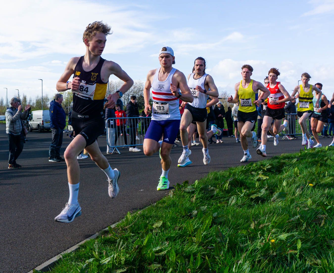 Male runners competing in a road race on a sunny day with spectators watching nearby.