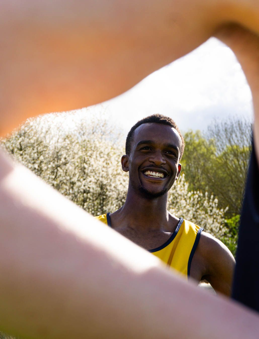 Smiling male athlete in yellow vest photographed through teammates arms at an outdoor running event.