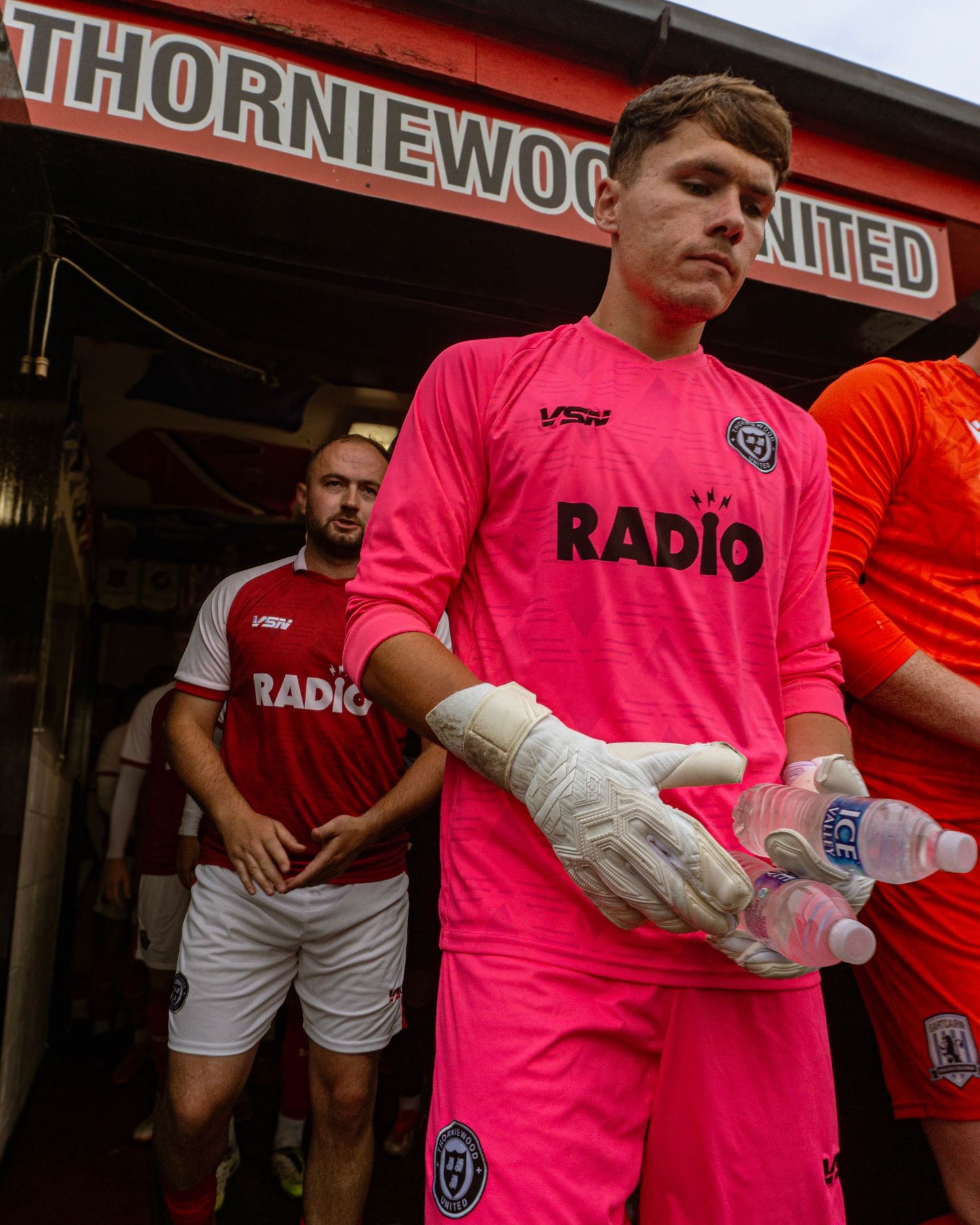 A goalkeeper in a pink kit with Radio sponsor exits tunnel at Thorniewood United football ground carrying gloves and water.