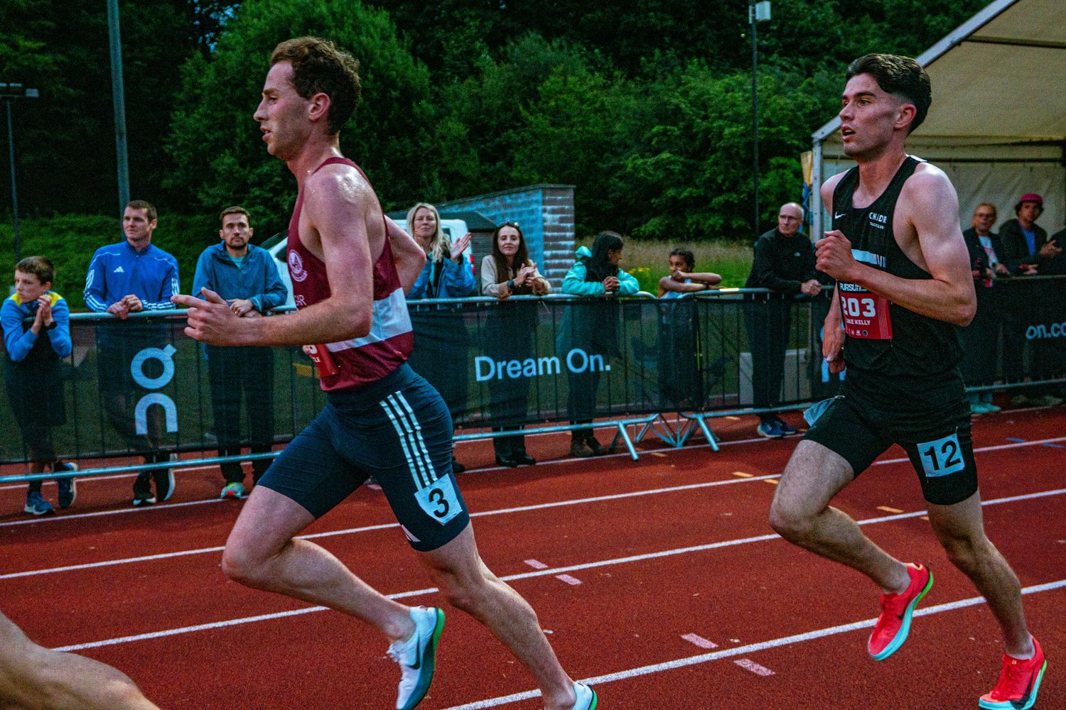 Two male athletes compete in a track race at an outdoor stadium with a Dream On banner in the background.