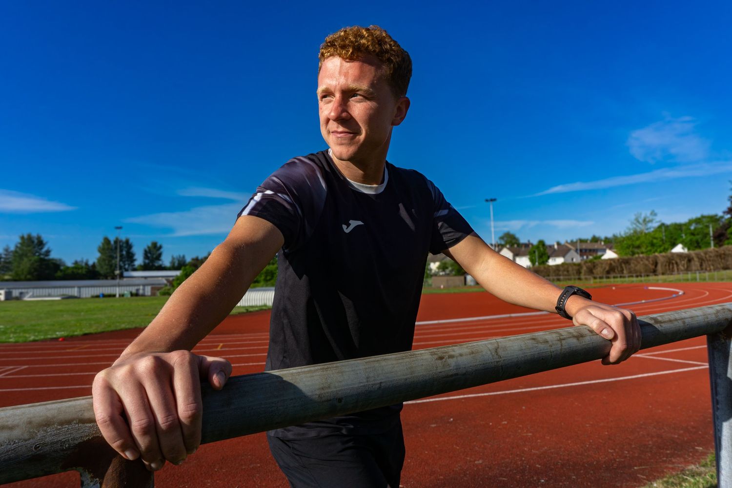 Young male runner leaning on a railing at an outdoor athletics track on a sunny day wearing a Joma shirt.