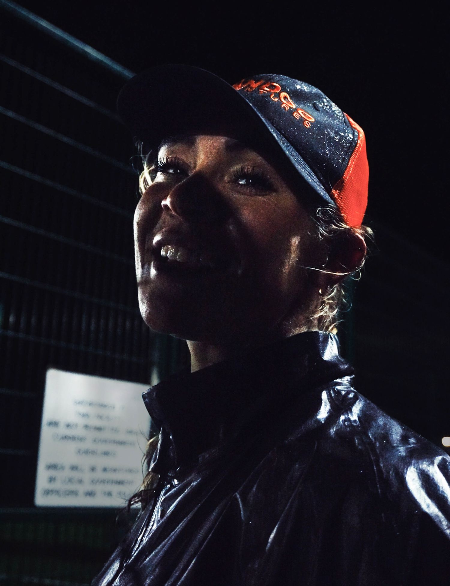Smiling woman in a wet rain jacket and orange cap under dark nighttime conditions outdoors.