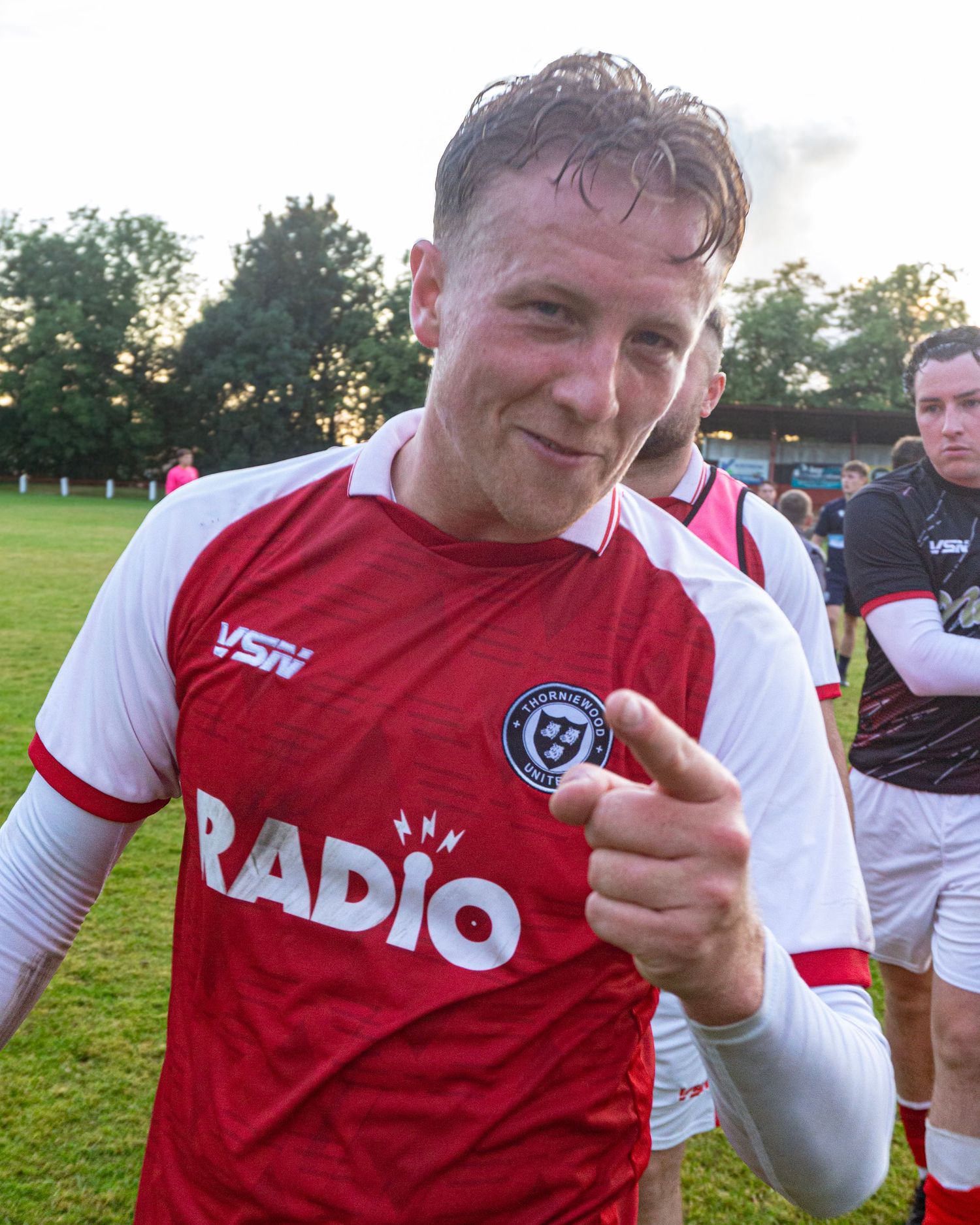 A footballer in a red and white Radio jersey smiles and points at the camera after a match.