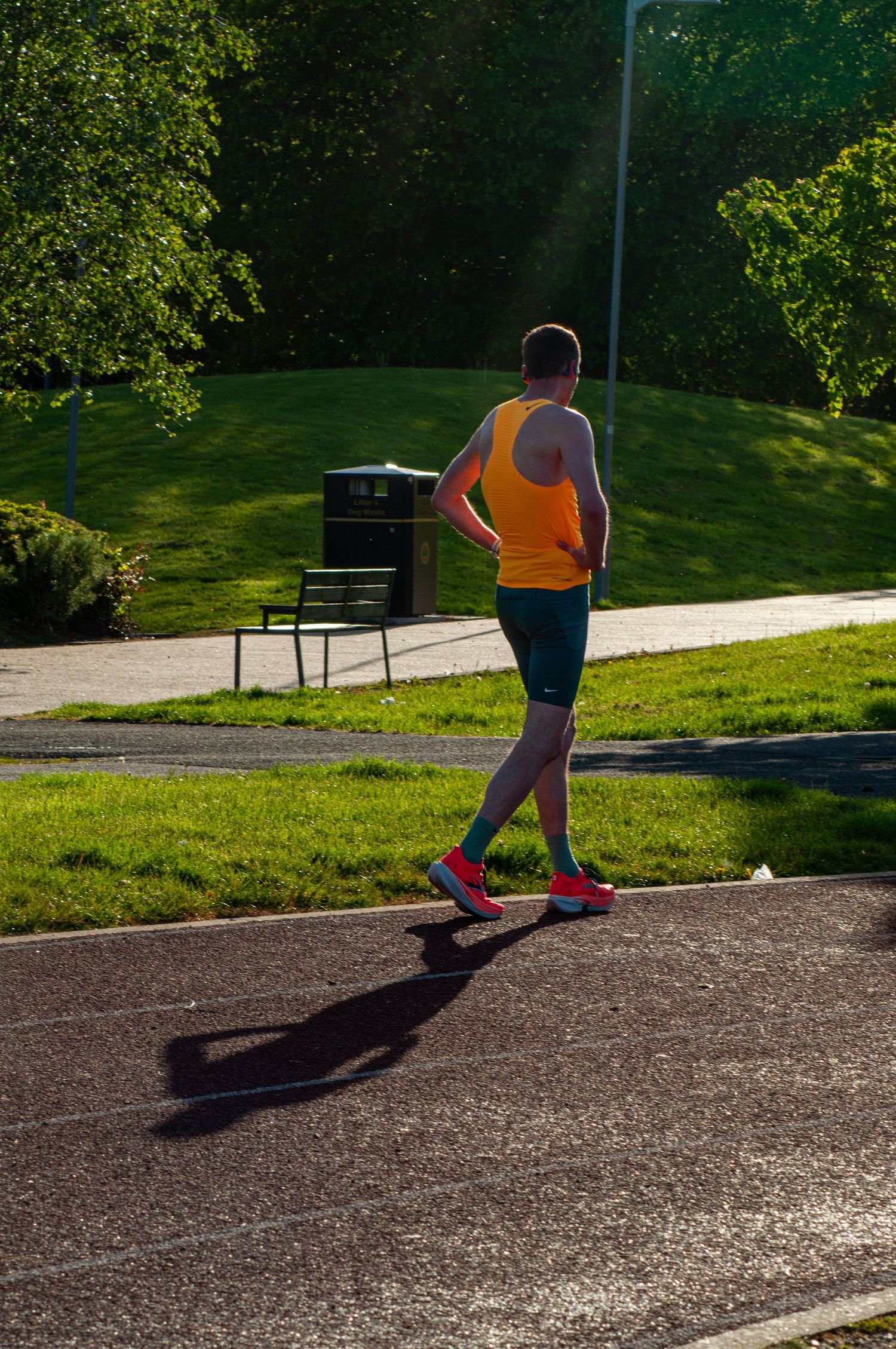 Male runner in orange vest and teal shorts jogging on a track beside green parkland.