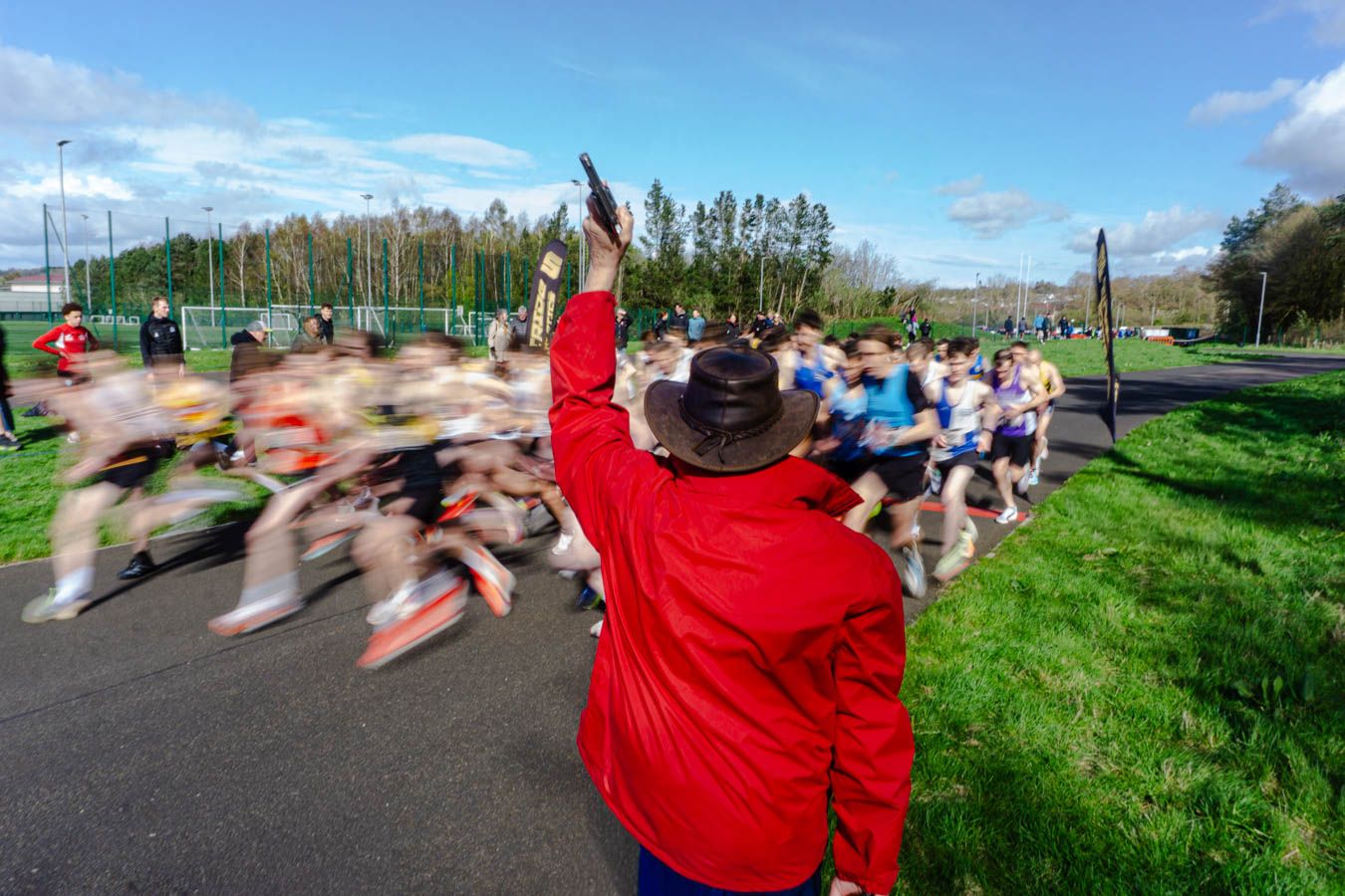 Race starter in red jacket fires starting pistol as runners surge forward at an outdoor road race.