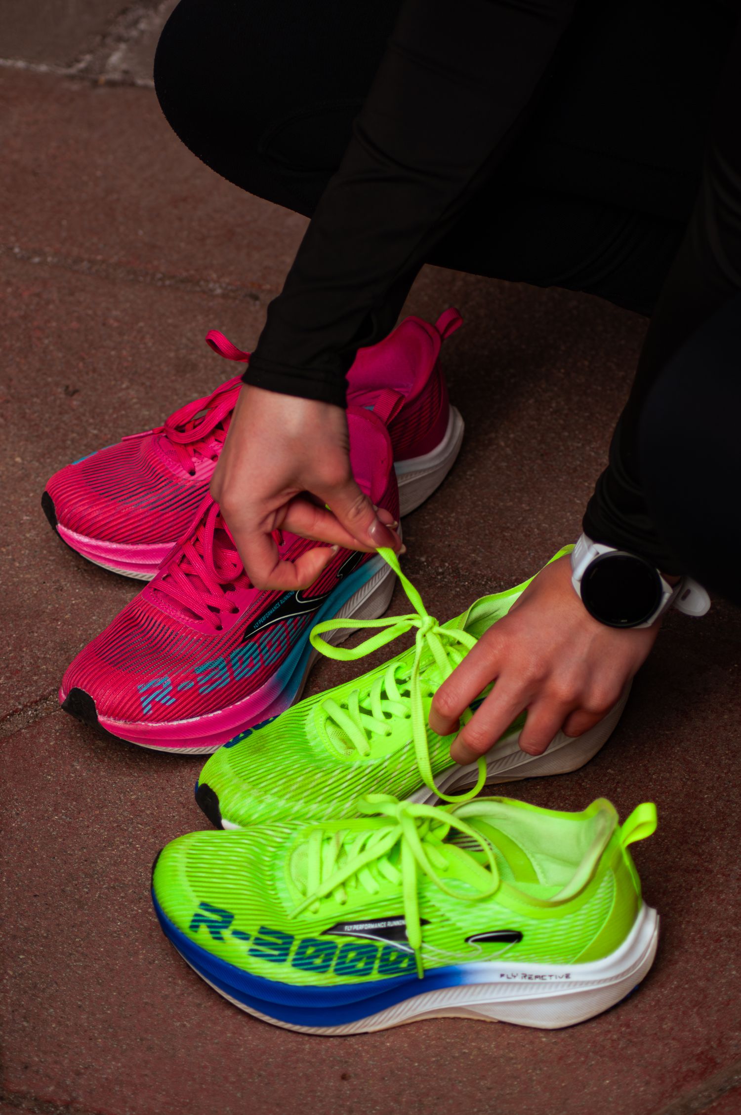 Close-up of hands tying neon green R-3000 running shoes next to pink sneakers with smartwatch visible.