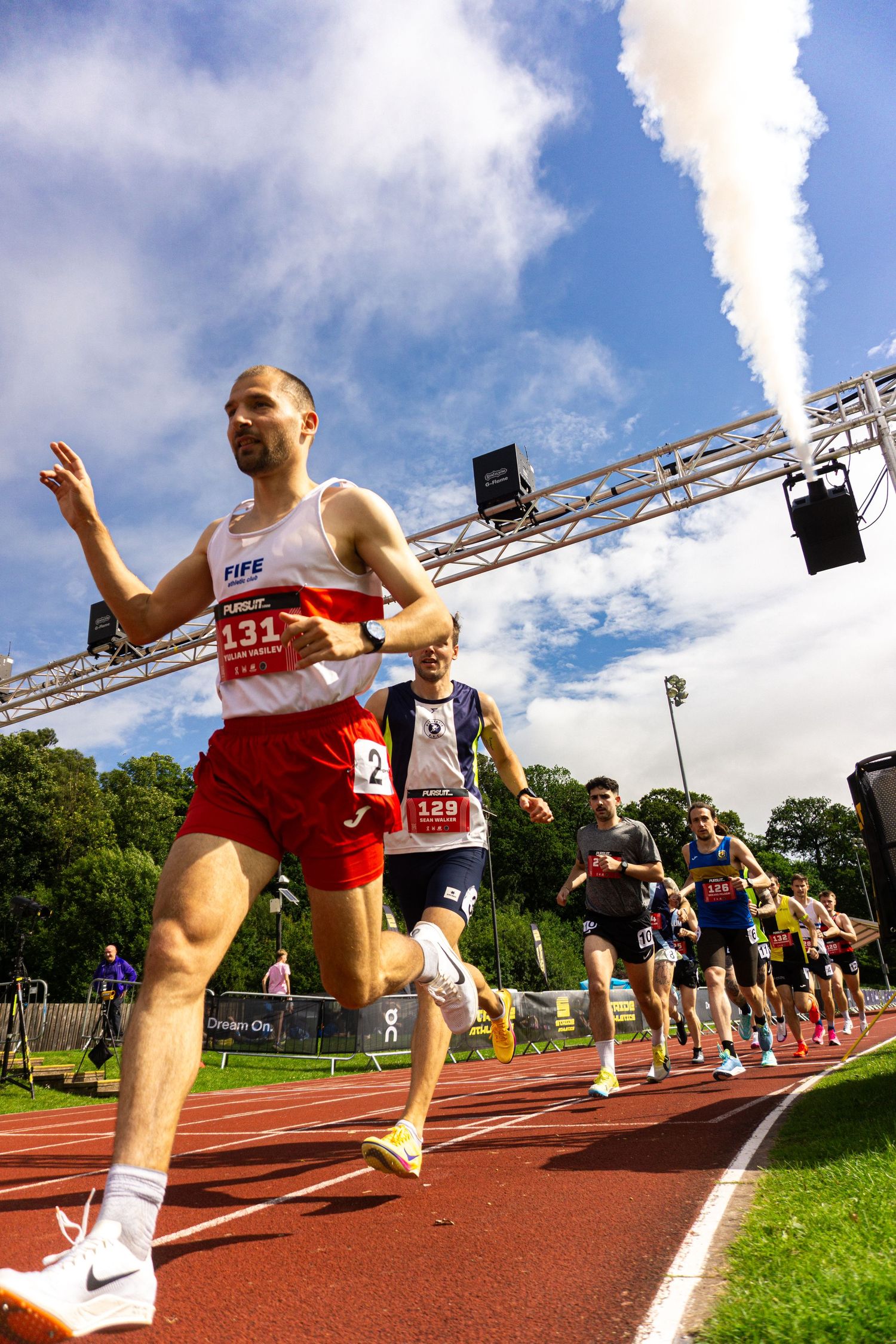 Runners compete on a red athletics track at a outdoor race event, led by athlete number 131 wearing a Fife vest.