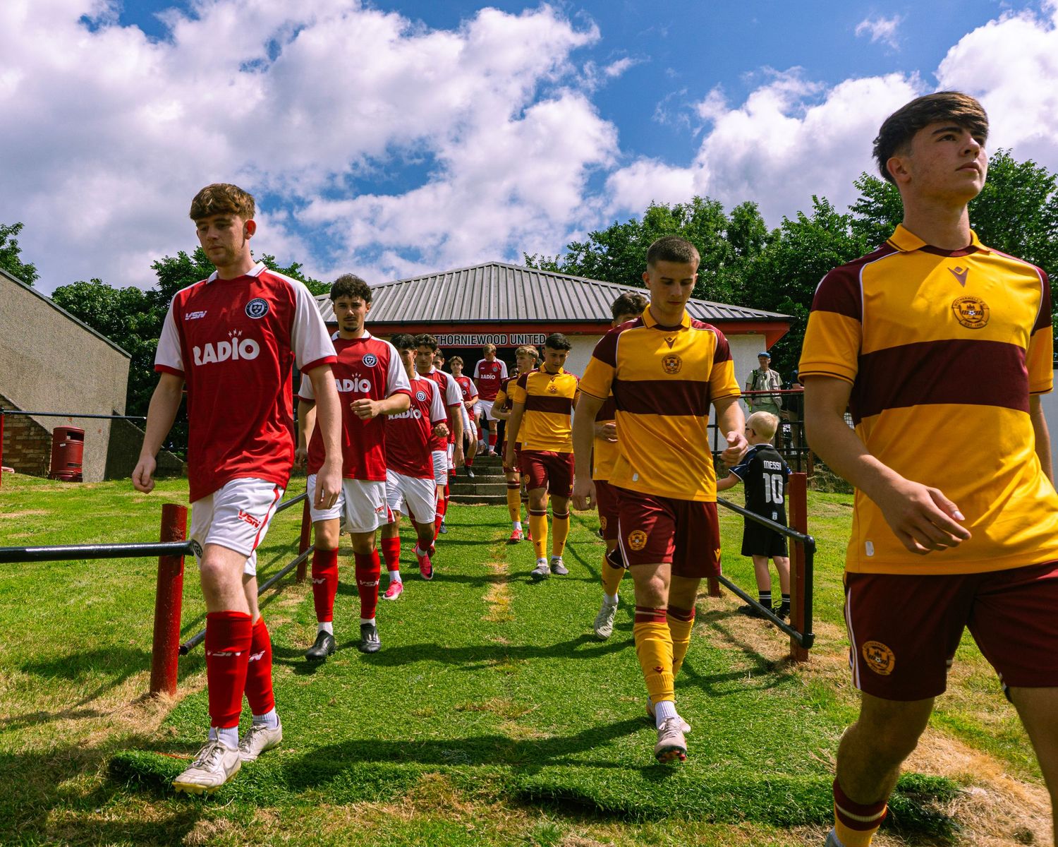 Young footballers in red-white and yellow-maroon kits walk out together before a sunny outdoor match.