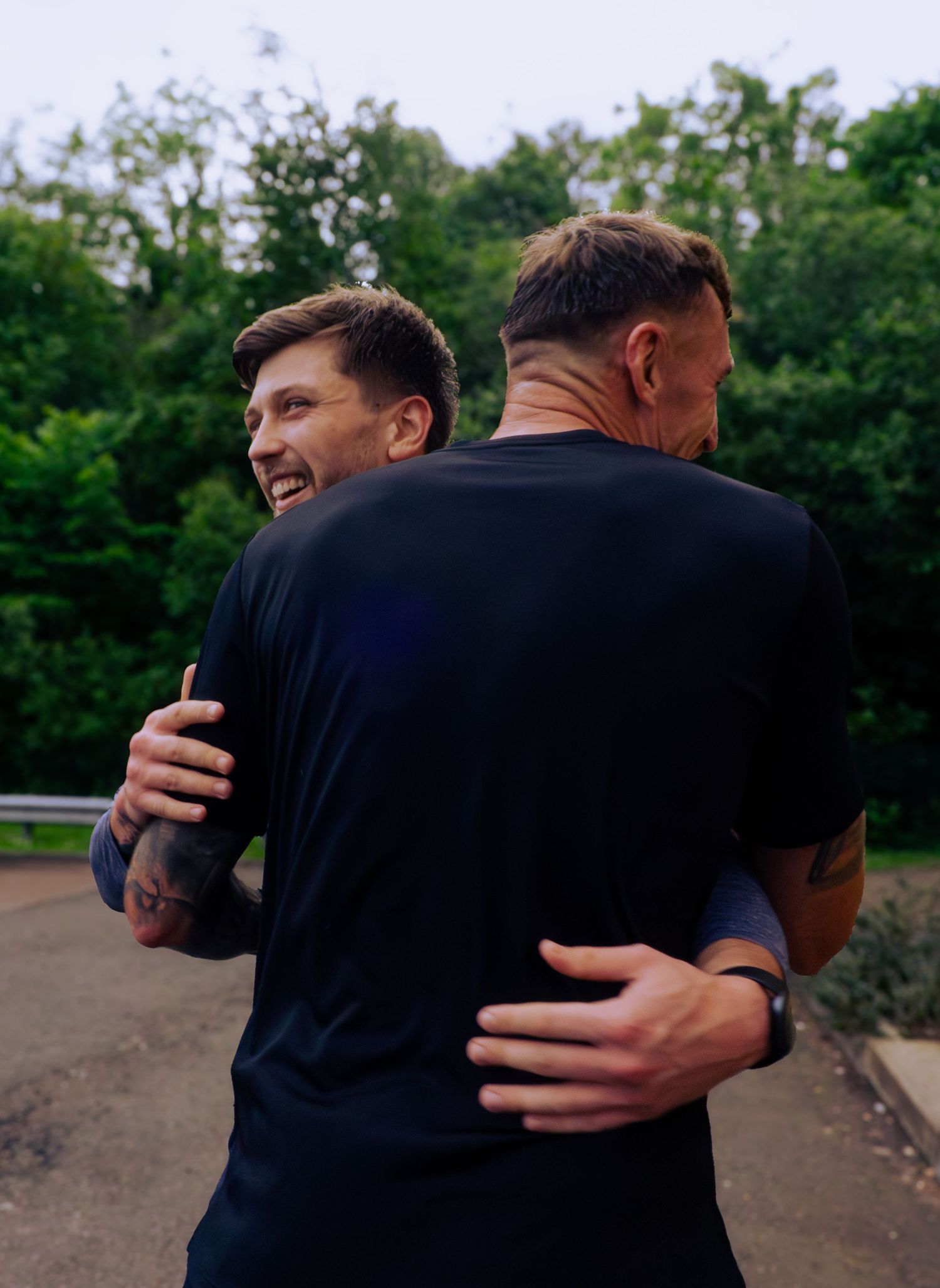 Two tattooed men in black t-shirts sharing a warm embrace outdoors with green trees in the background.