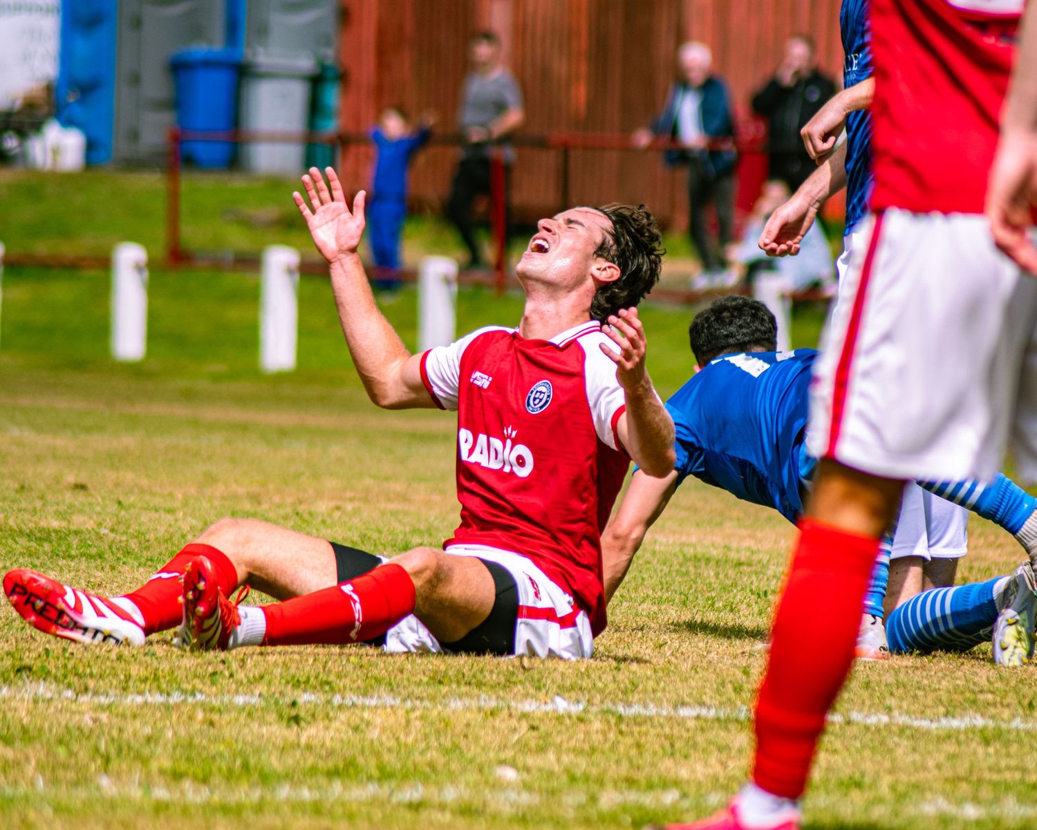 Soccer player in red kit appeals to referee after falling on grass during a competitive match.