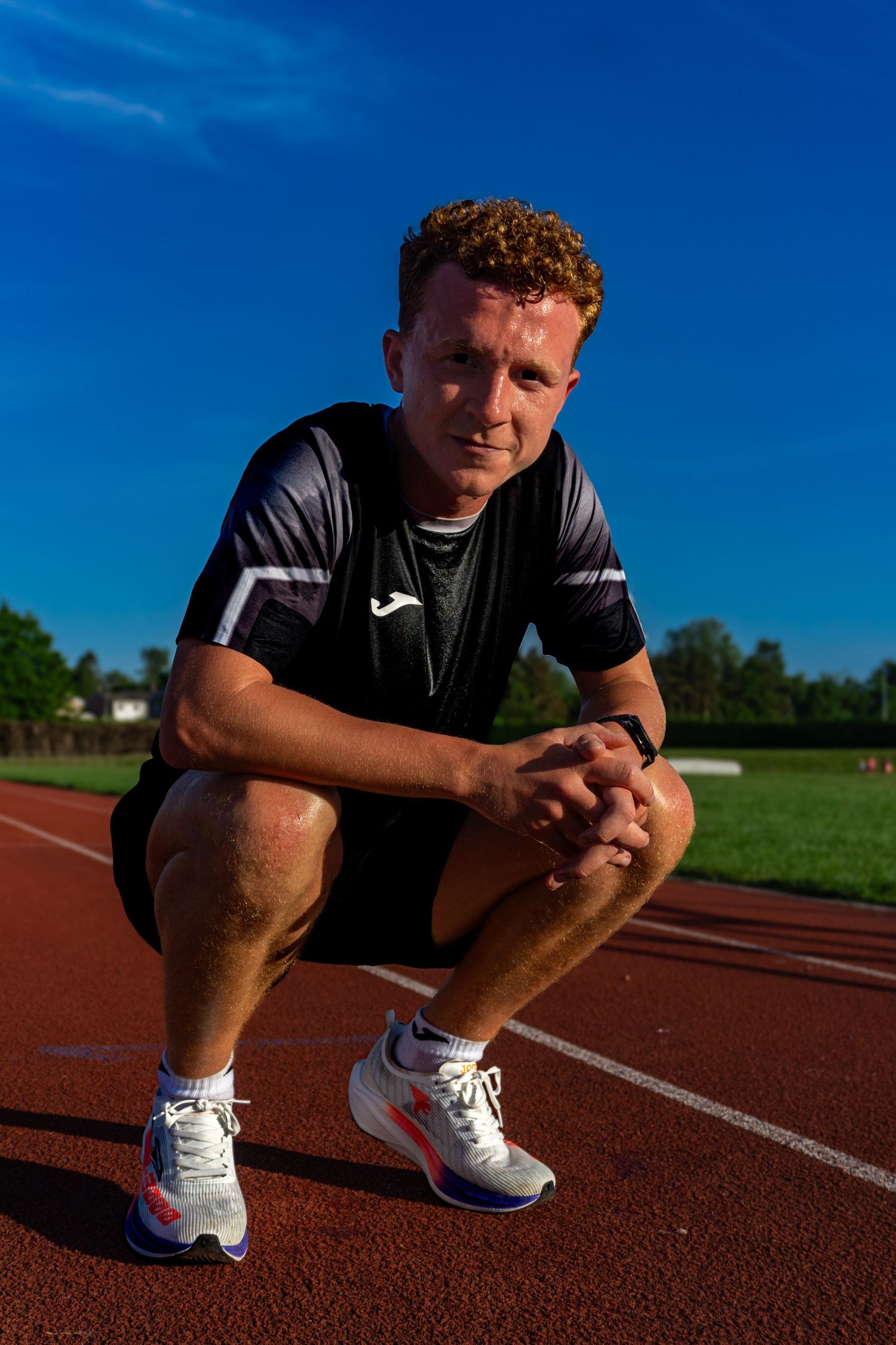 Male athlete crouching on a red running track in Joma sportswear and white running shoes, looking at camera.
