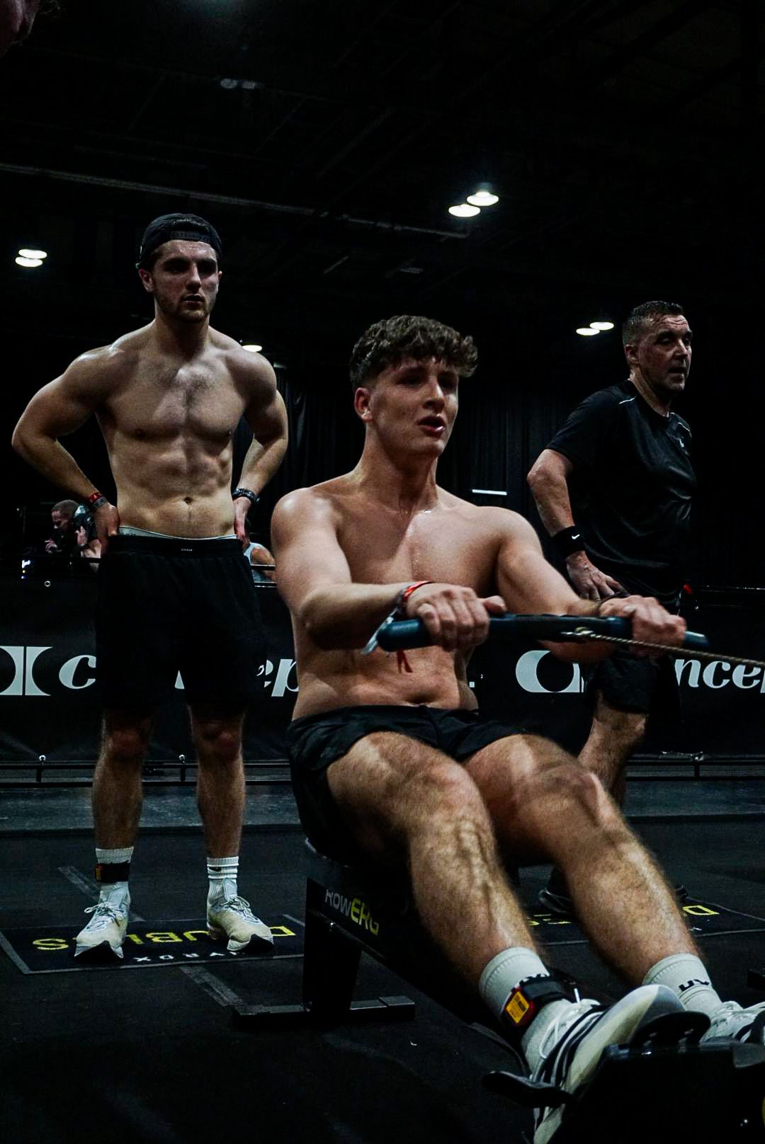 Focused male athlete rowing on a Concept2 machine while a teammate watches during a gym competition.
