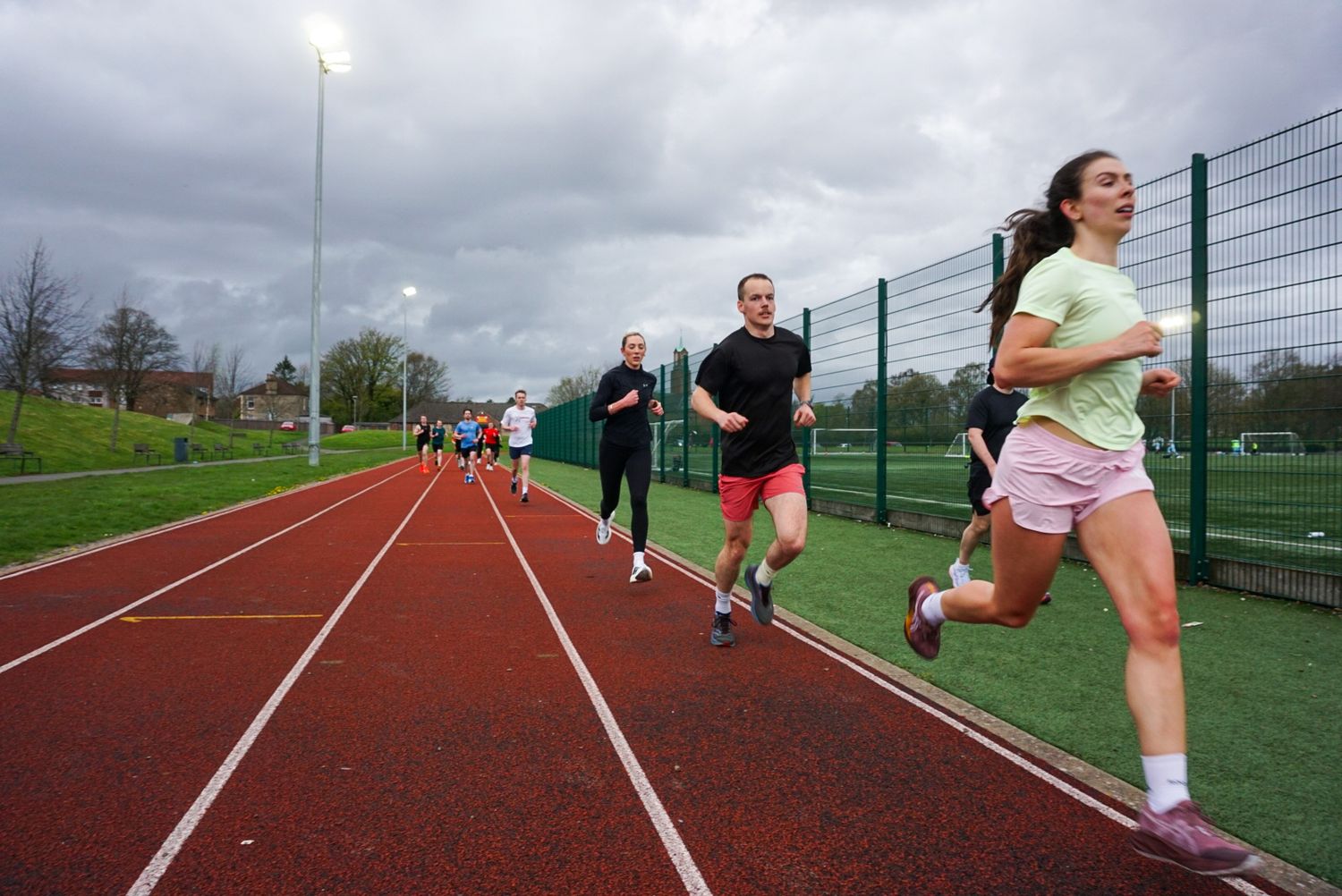 Group of runners training on a red outdoor athletics track under cloudy skies.