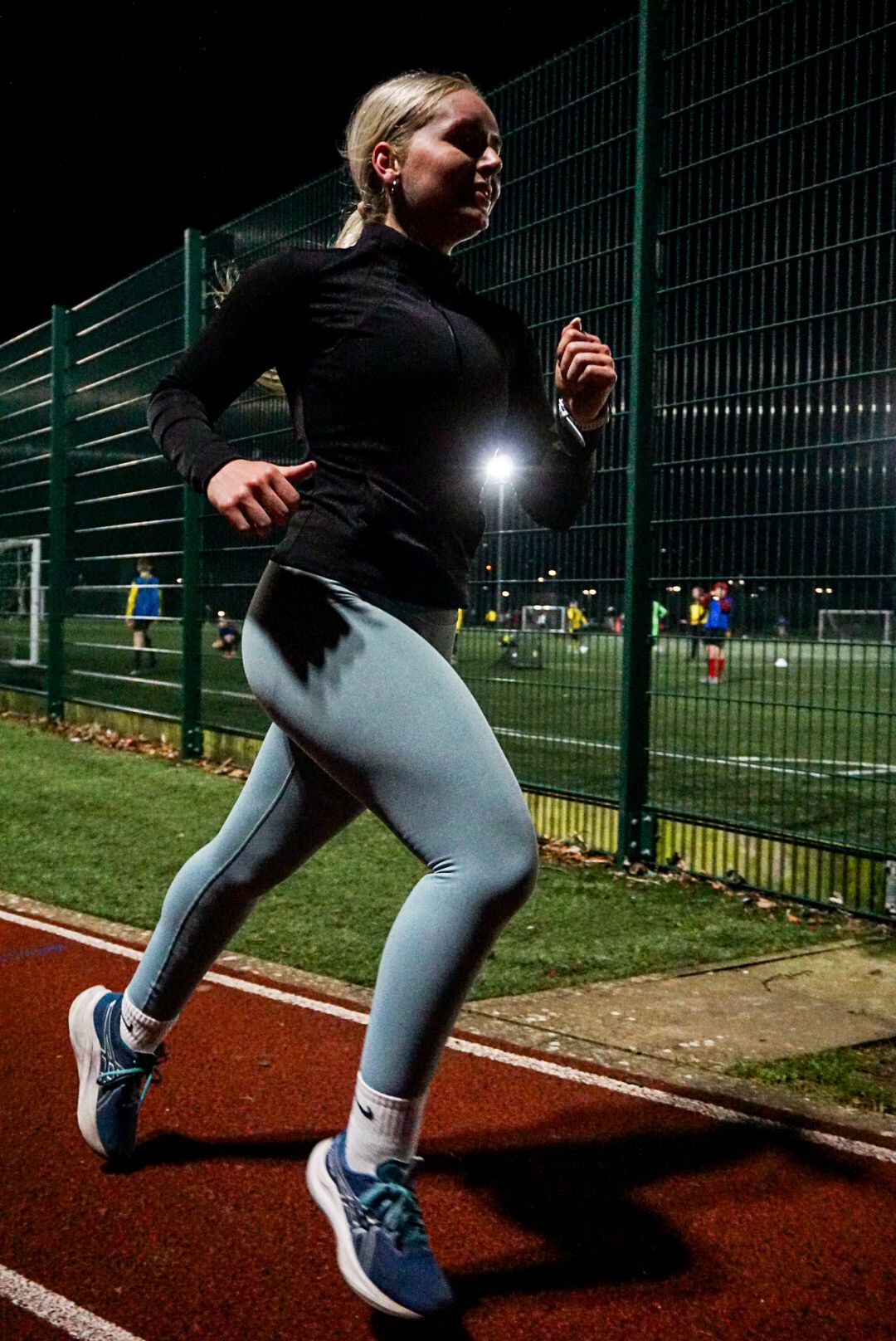 Blonde woman running on a red athletic track at night wearing light blue leggings and black jacket.