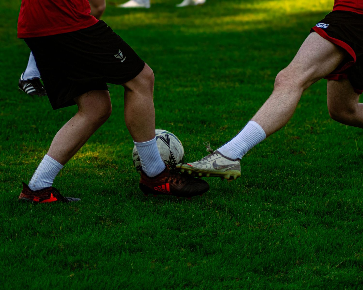 Two soccer players compete for the ball on a green grass field wearing cleats and black shorts.
