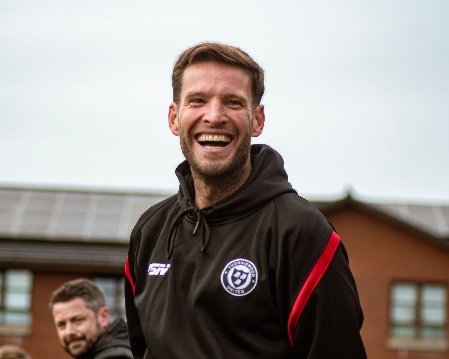 Smiling man in black and red sports hoodie laughing outdoors at a sporting event.