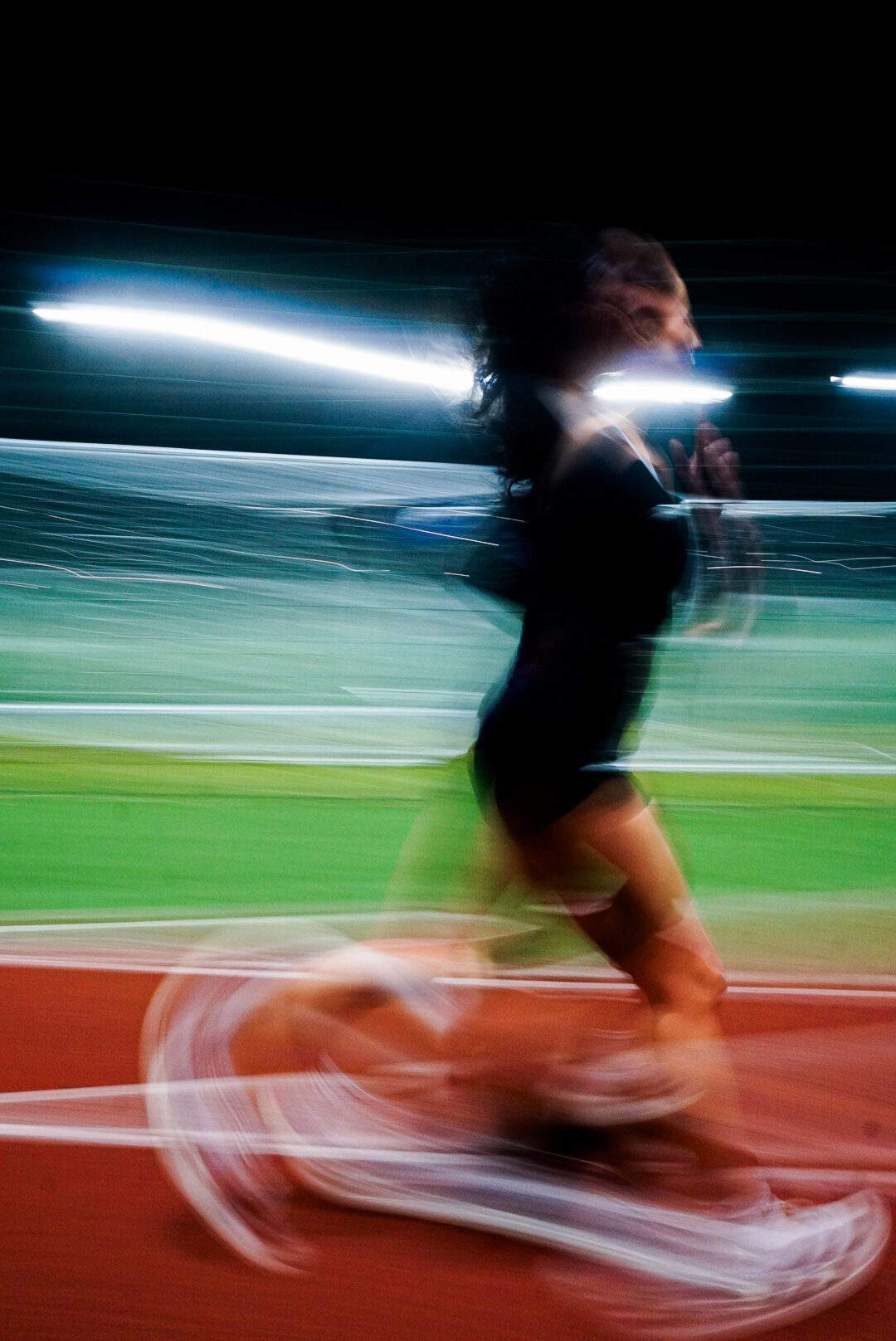 Motion blur photo of a curly-haired runner in black sprinting on a red athletic track at night.