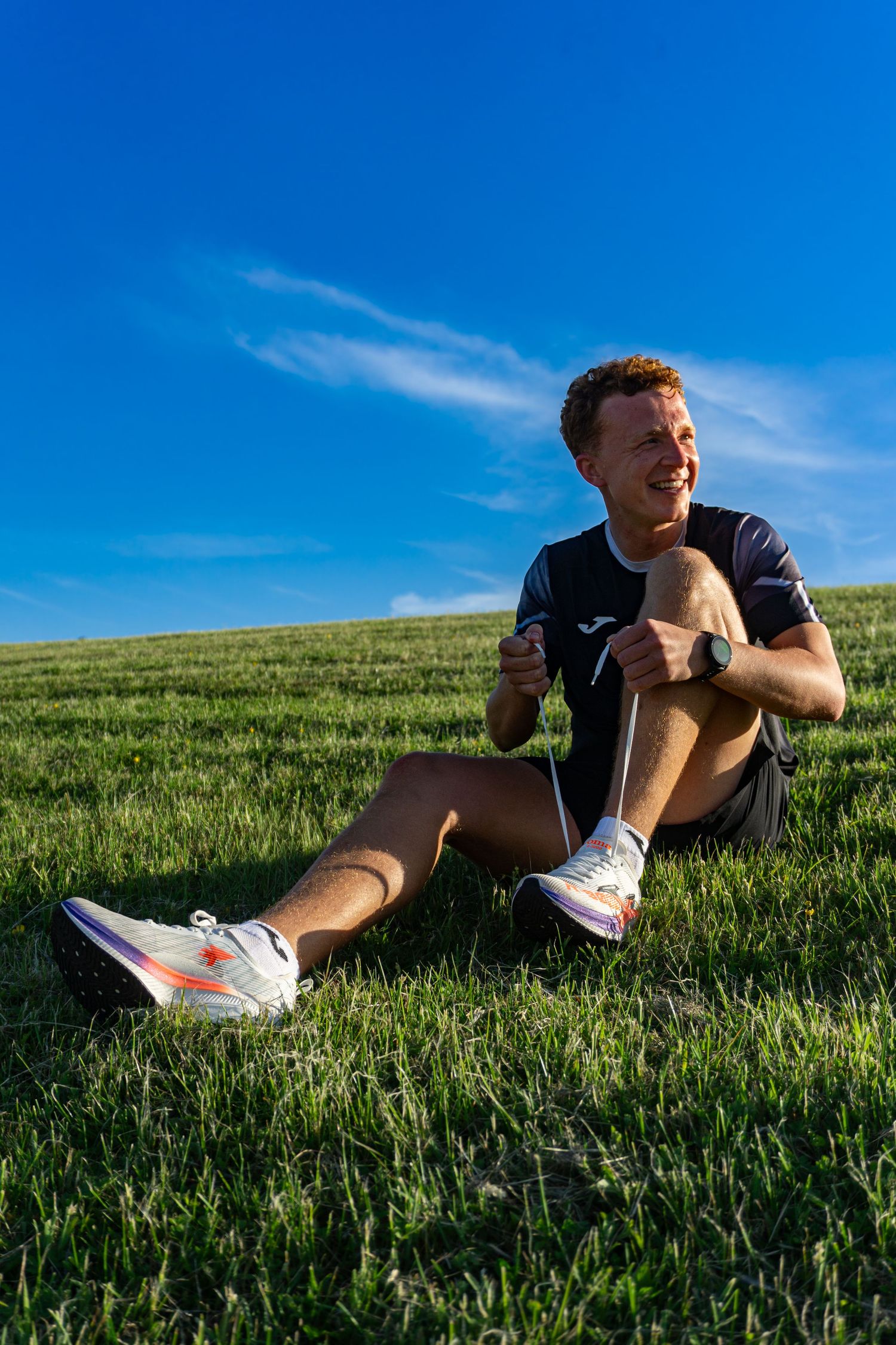 Smiling male runner sitting on green grass wearing Joma gear and athletic shoes under a bright blue sky.