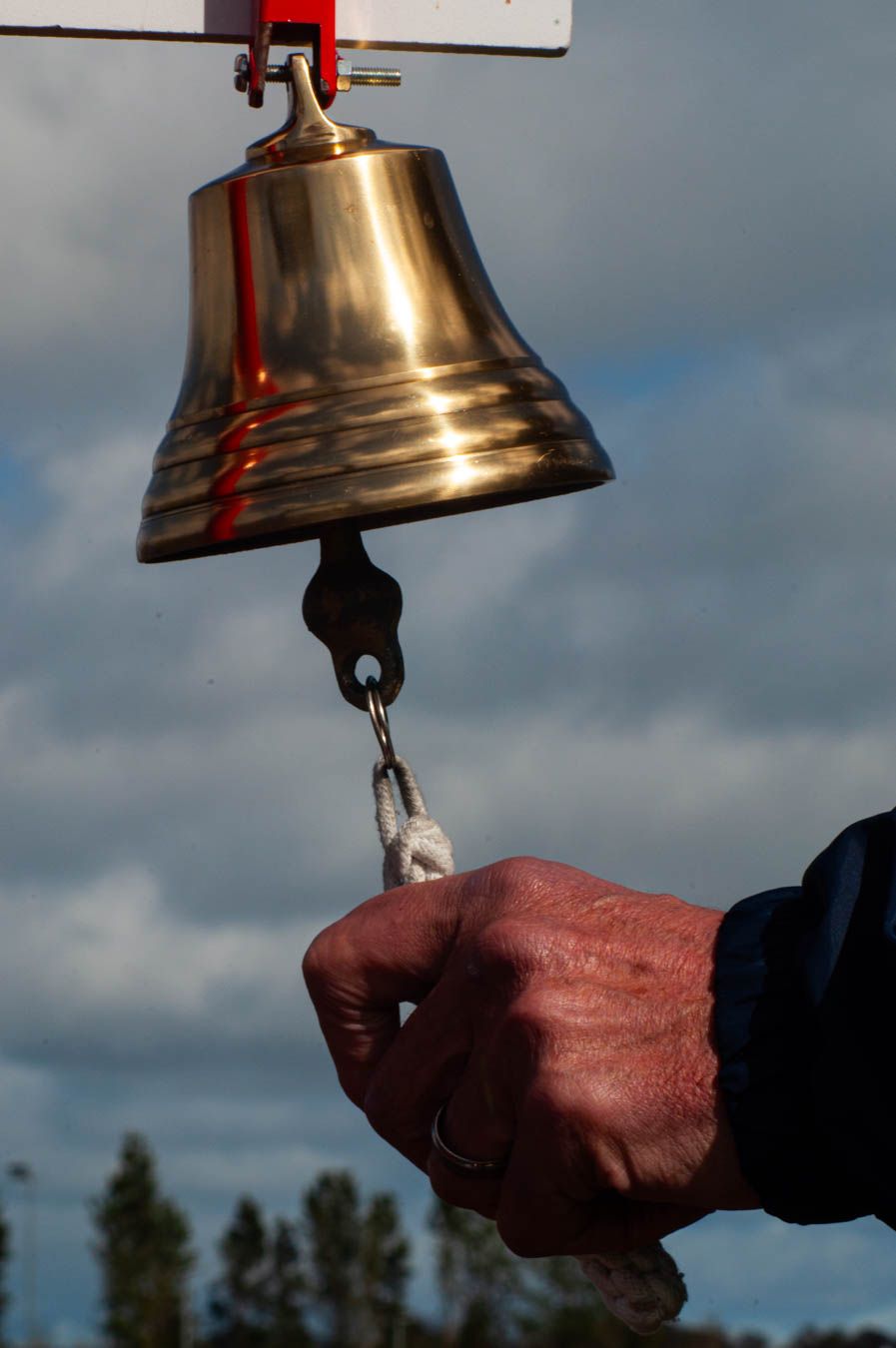Hand ringing a brass bell at outdoor sporting event against cloudy sky.