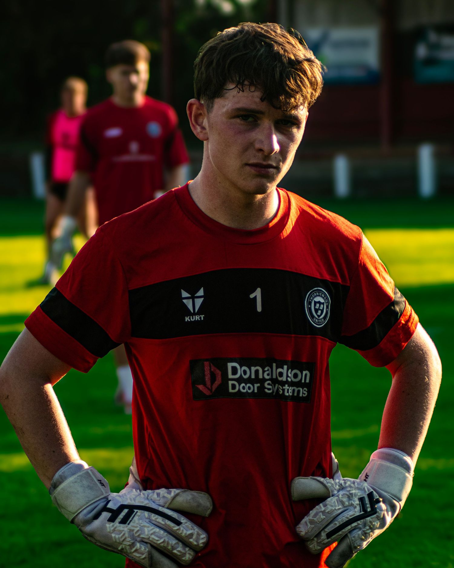 Young goalkeeper in red jersey numbered 1 with Donaldson Door Systems sponsor stands on green football pitch.