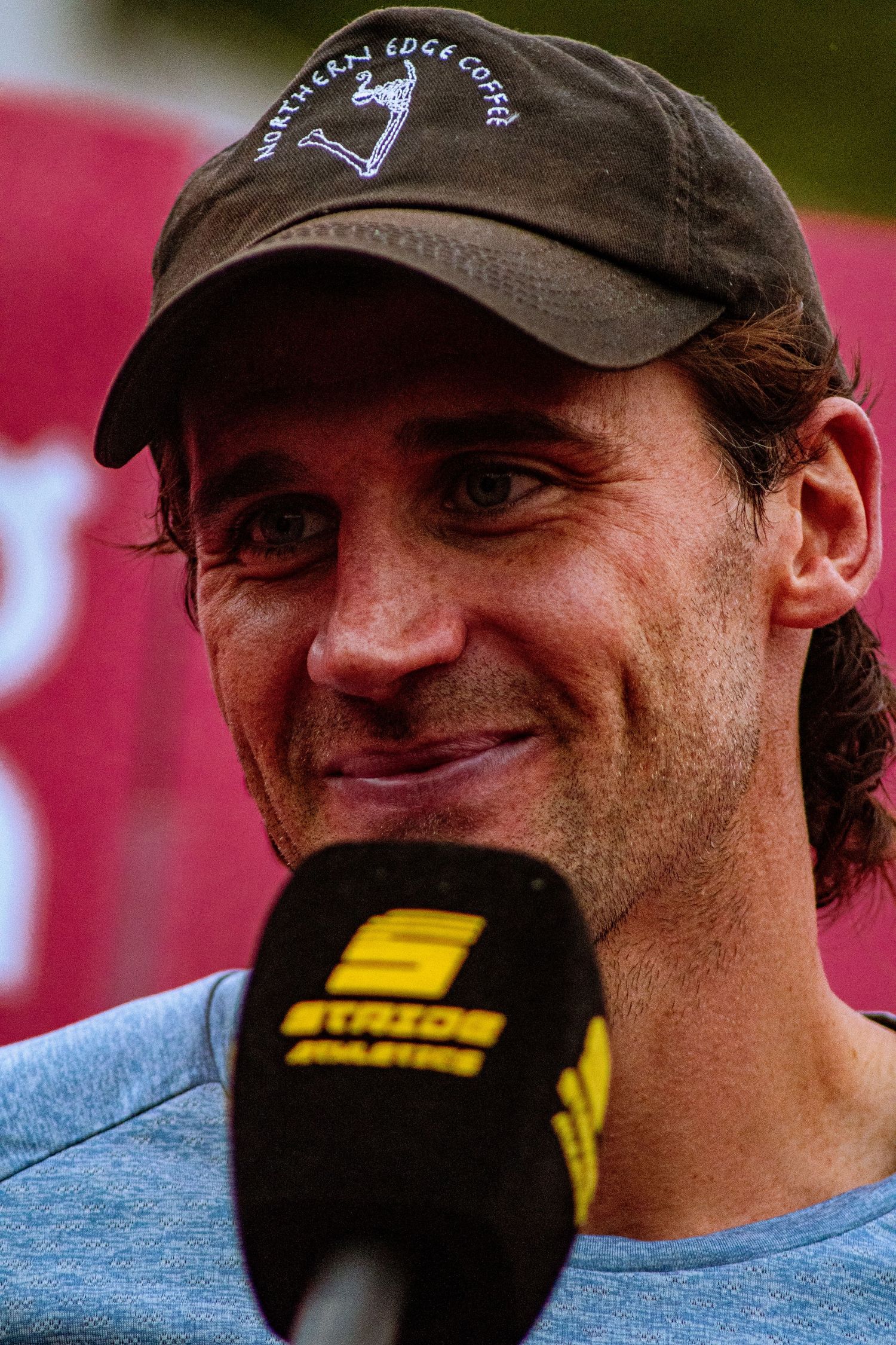Smiling man wearing a dark baseball cap and blue t-shirt being interviewed with a Stride Athletics microphone outdoors.