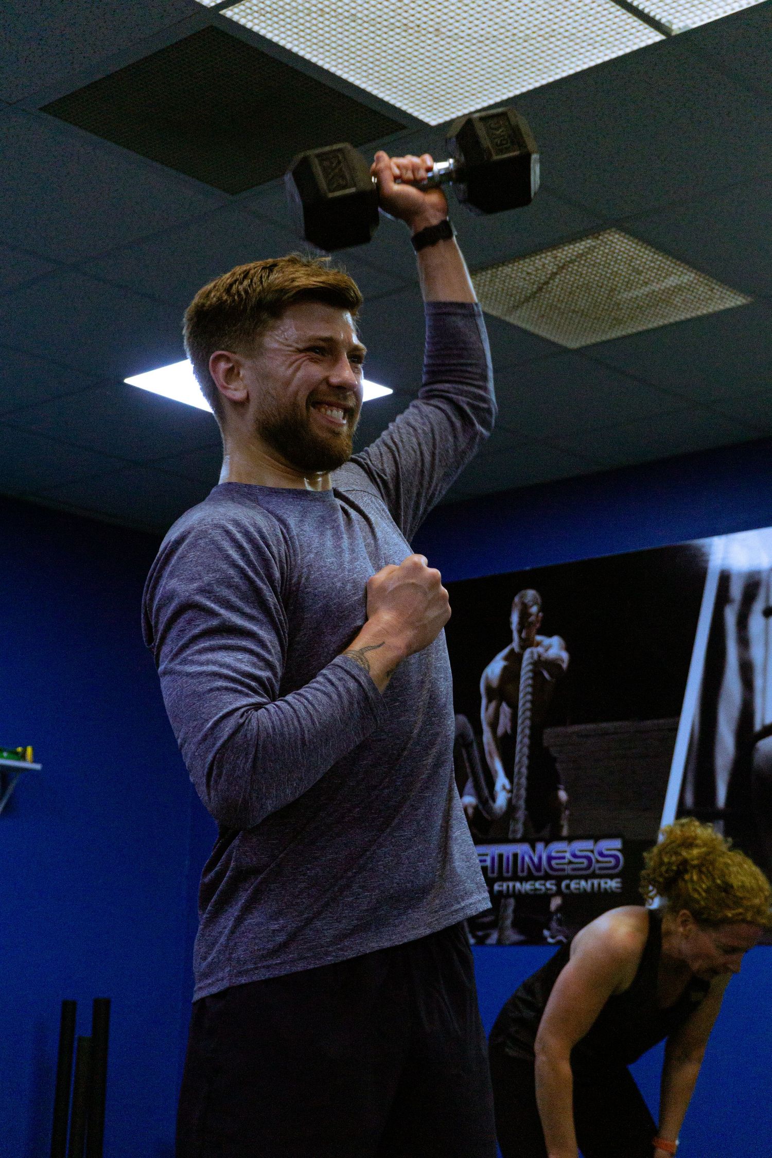 Smiling man lifting a dumbbell overhead with one arm in a blue gym during a workout session.