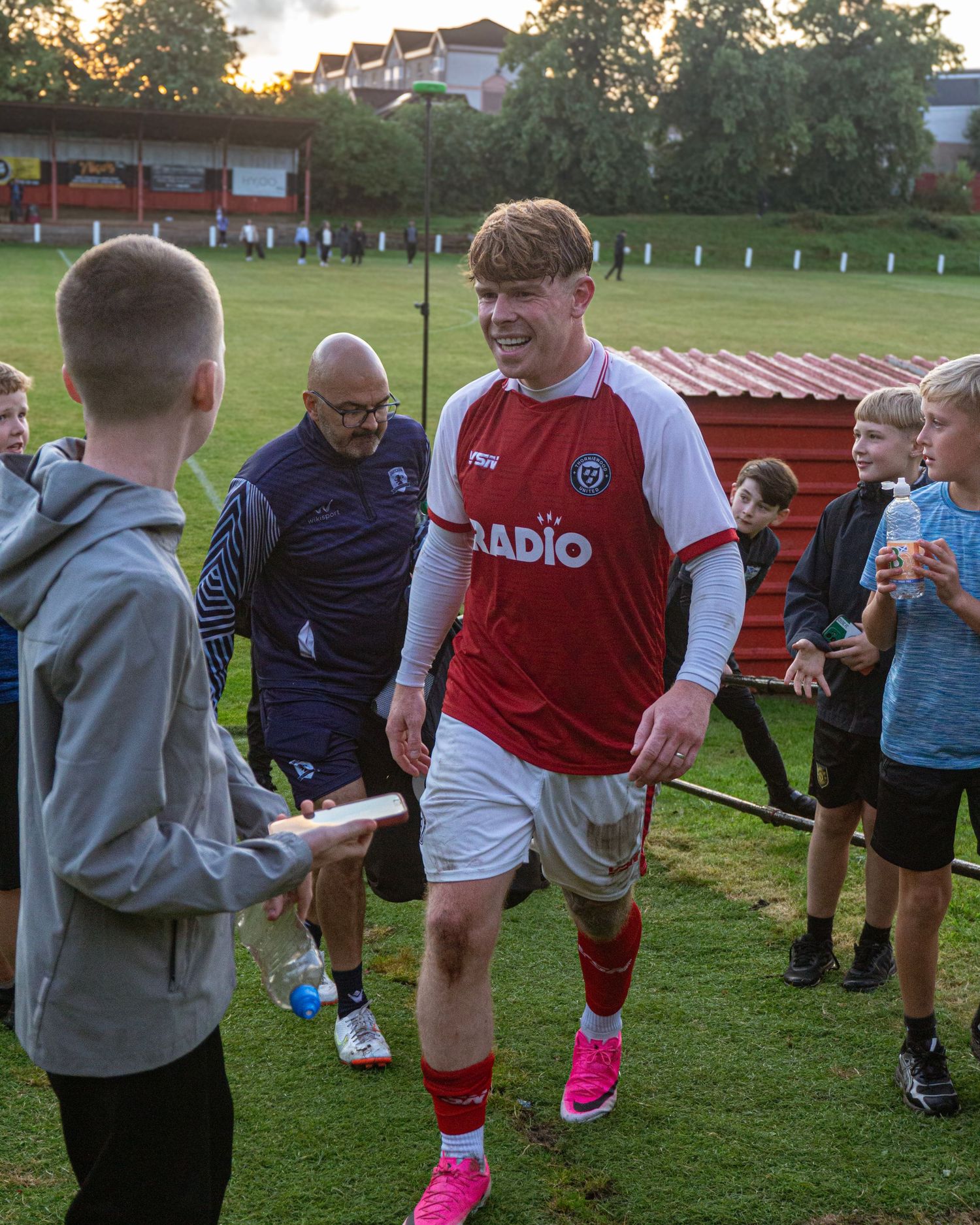 A young footballer in a red and white Radio kit greets fans on the pitch after a game.