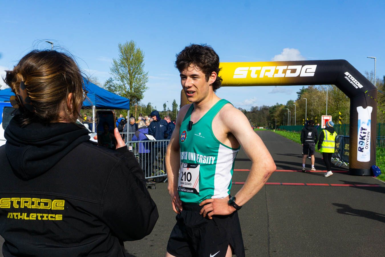 Male runner in green vest speaks to a photographer at the Stride Athletics finish line after a race.