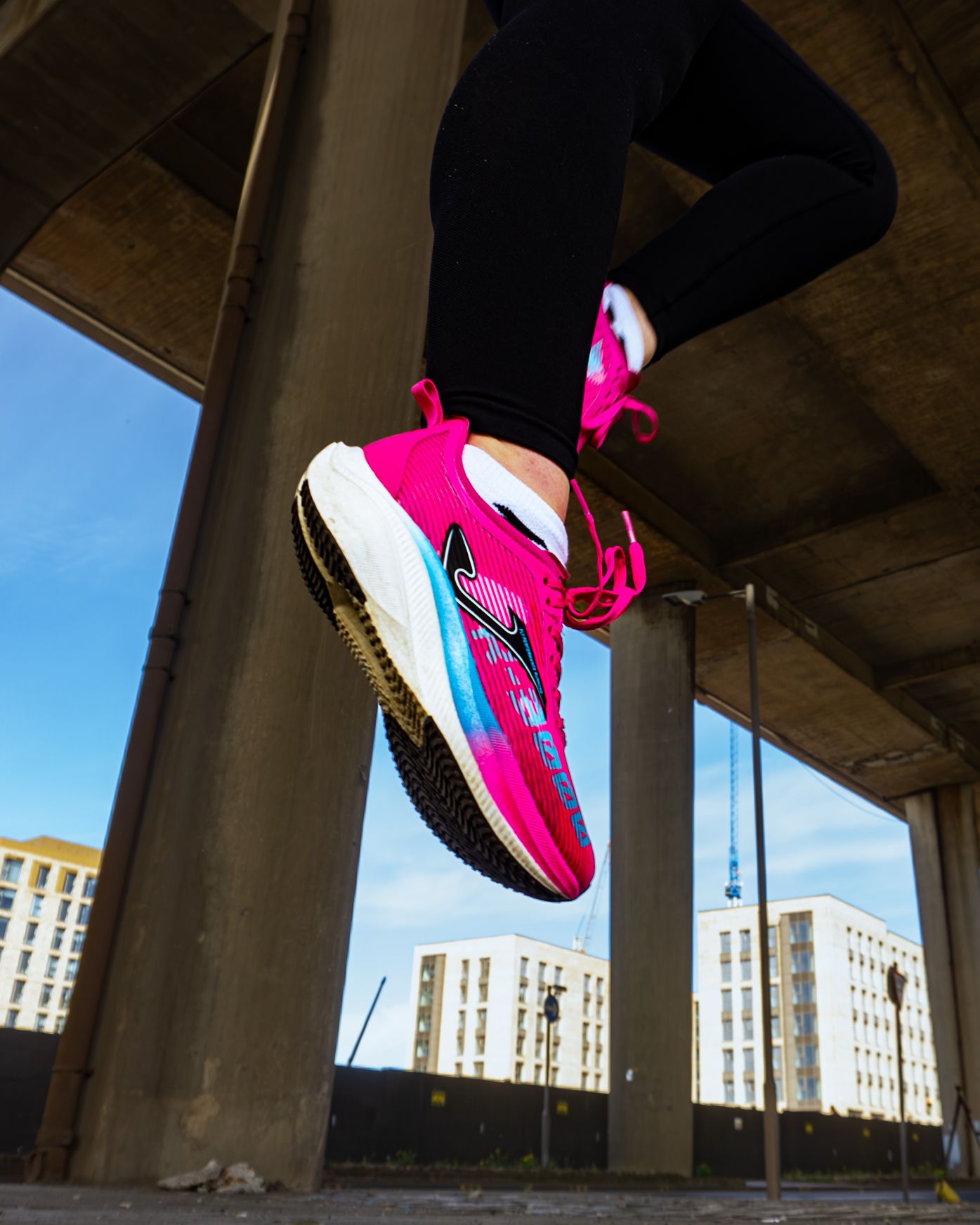Low angle shot of vibrant pink Joma running shoes worn by athlete under urban bridge structure.