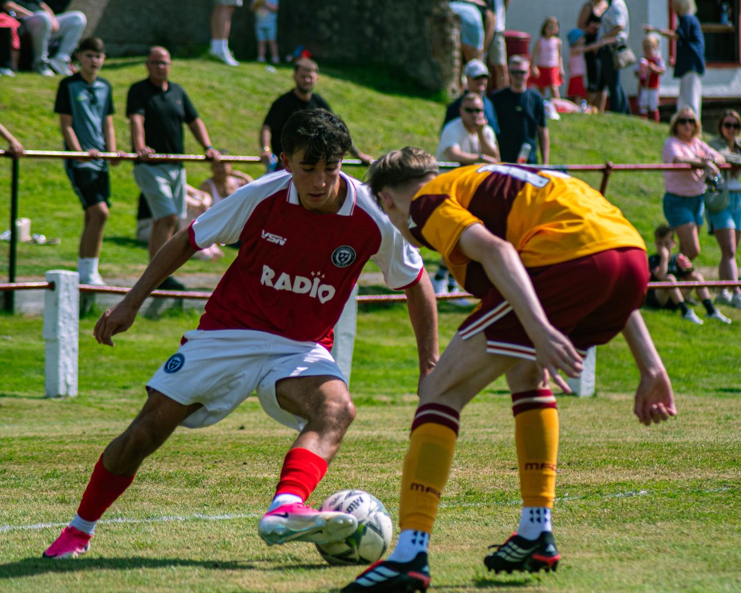 Two soccer players compete for the ball during a sunny outdoor match, one in red and white, the other in yellow and maroon.