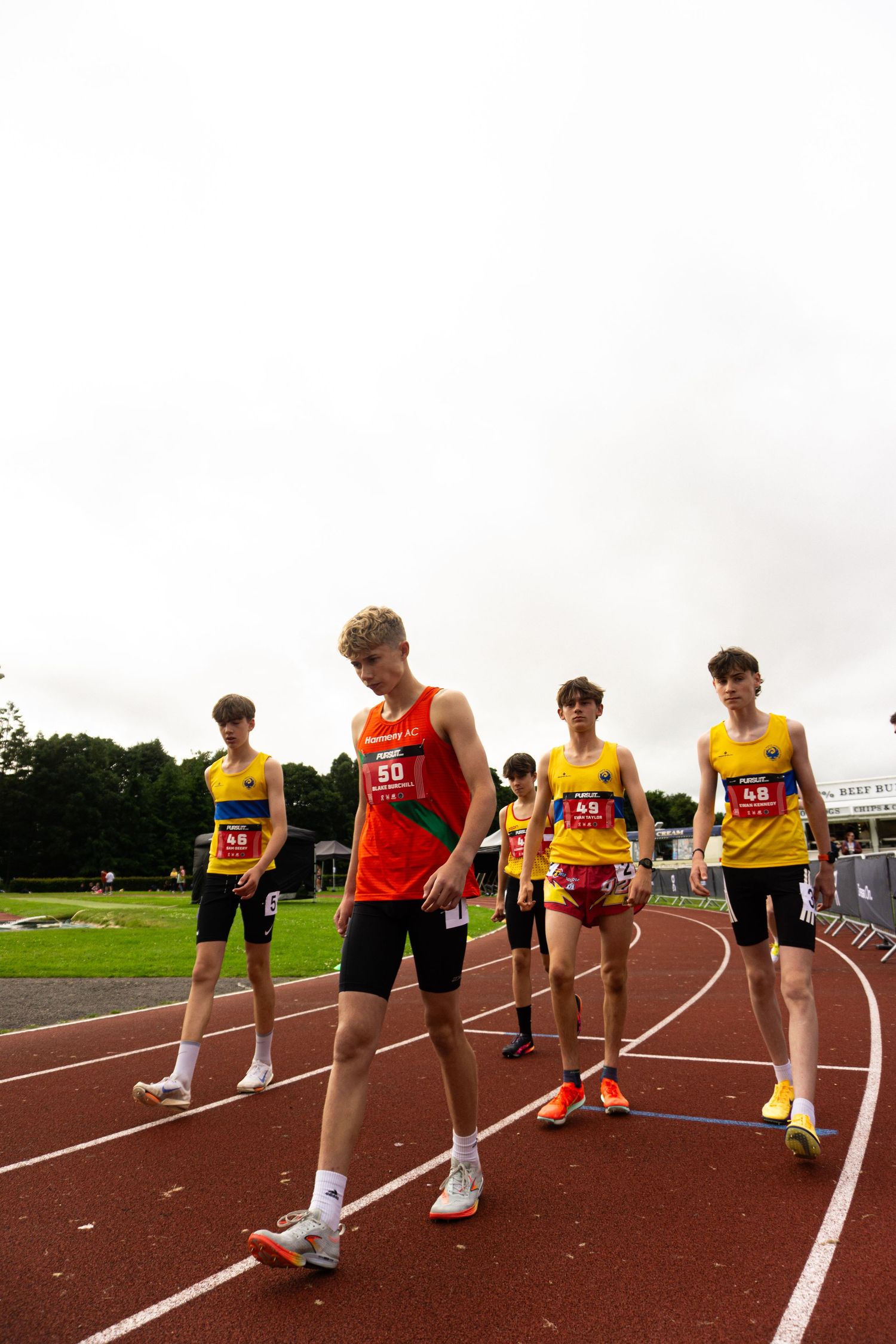 Young male athletes in numbered bibs prepare on a red running track at an outdoor athletics competition.