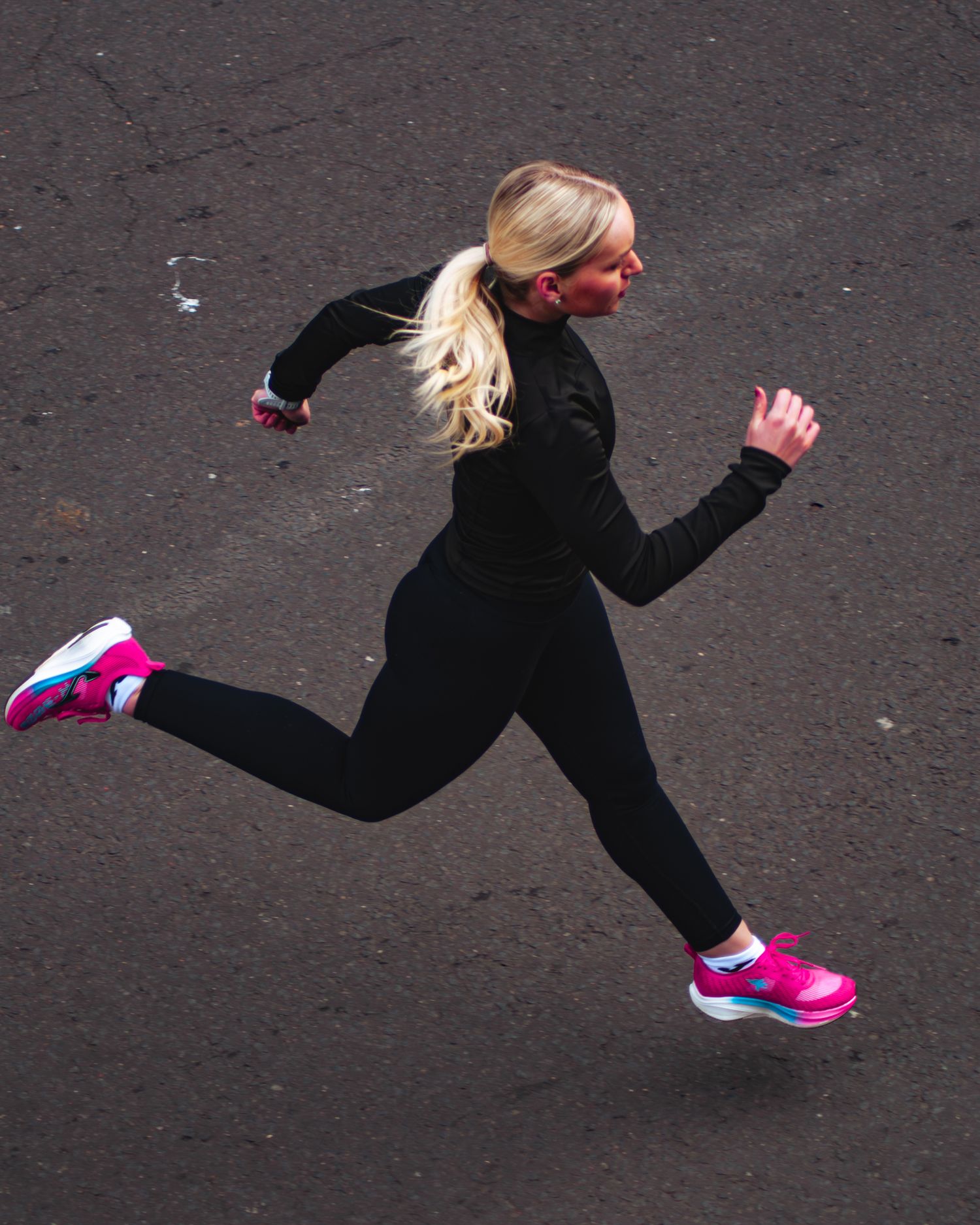 A blonde female runner in all-black athletic wear and pink Joma sneakers sprints across a dark asphalt surface.
