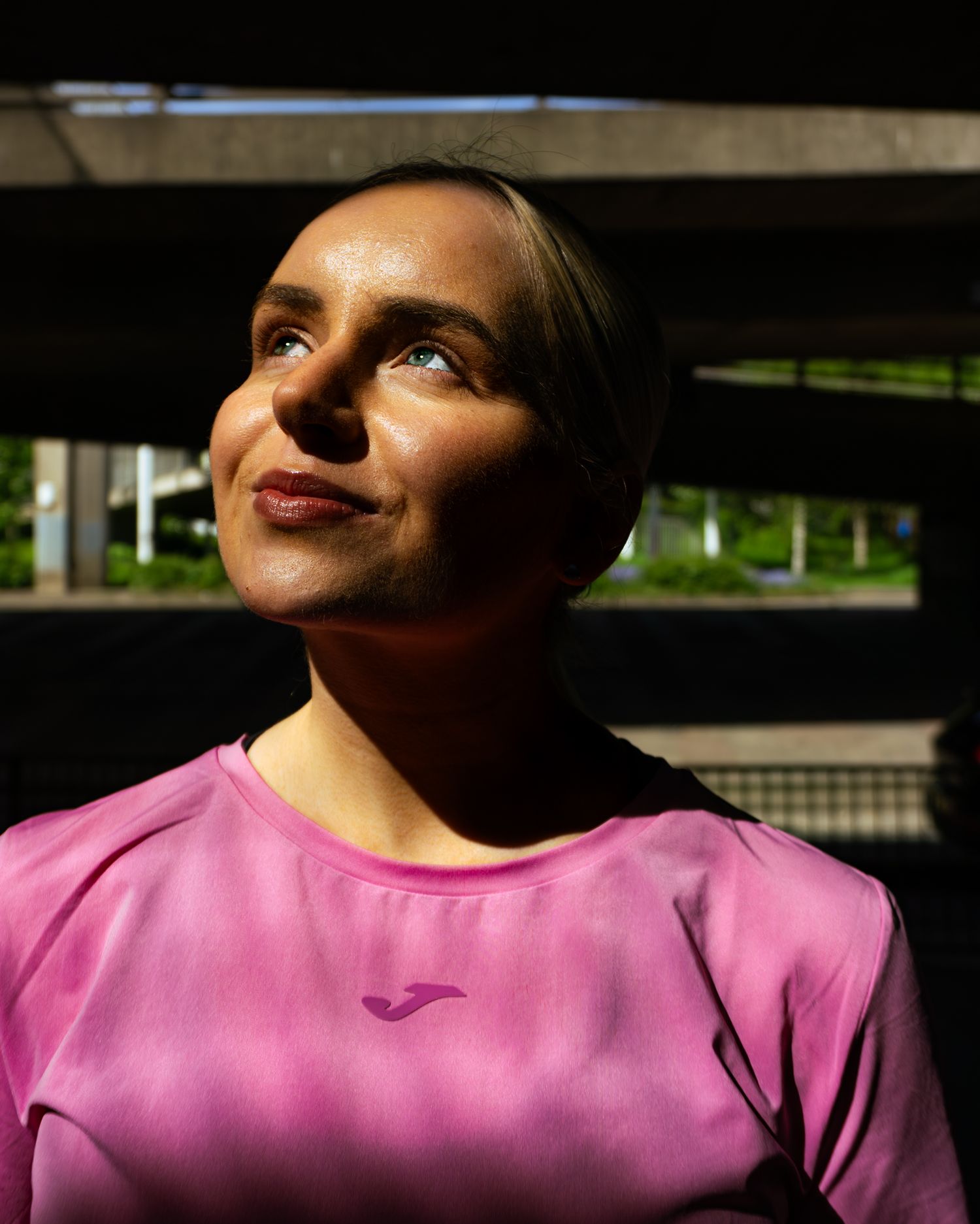 Close-up portrait of woman in pink Joma athletic shirt looking upward in dappled sunlight.
