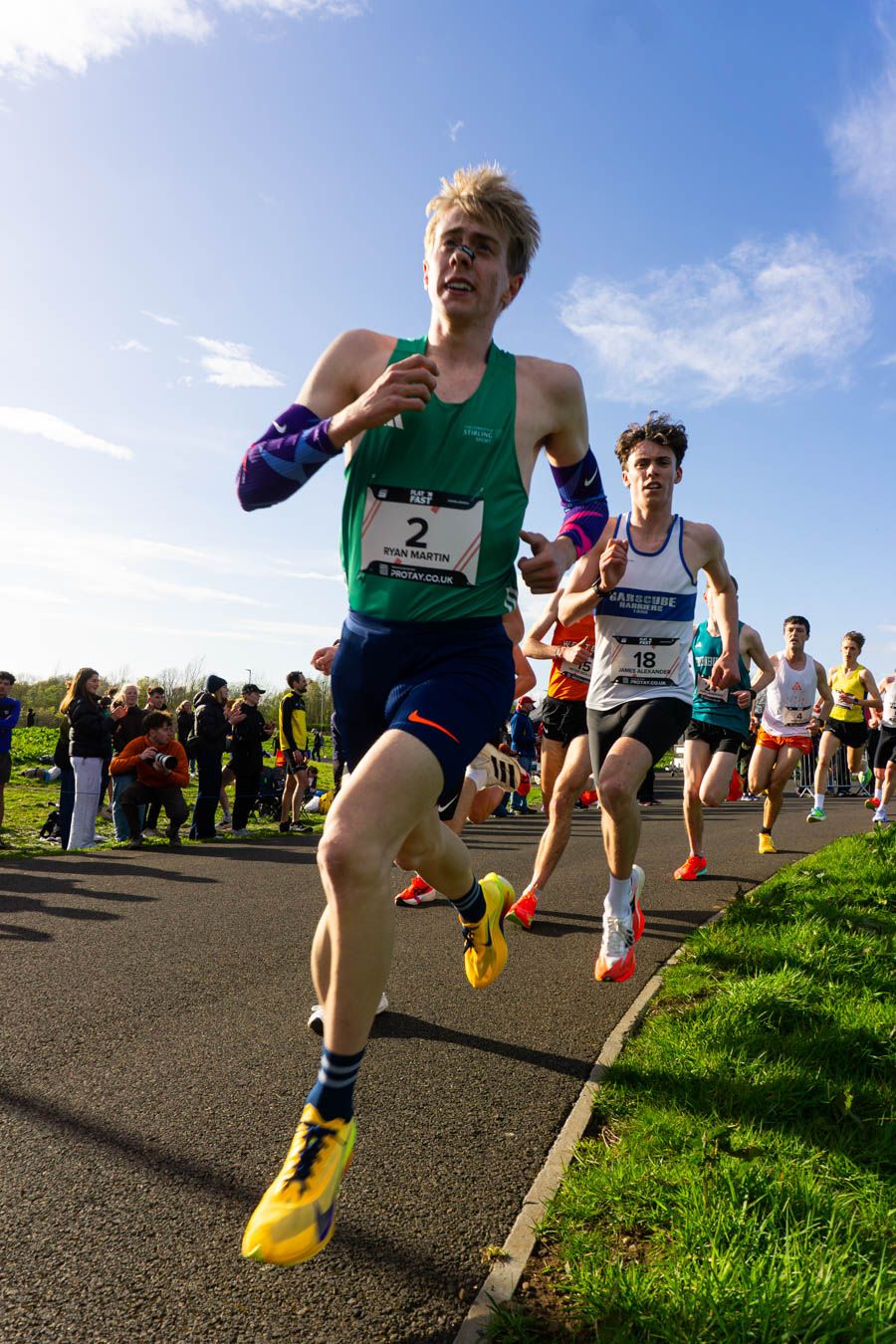 Runner wearing bib number 2 leads a road race in a green vest and yellow shoes on a sunny day.