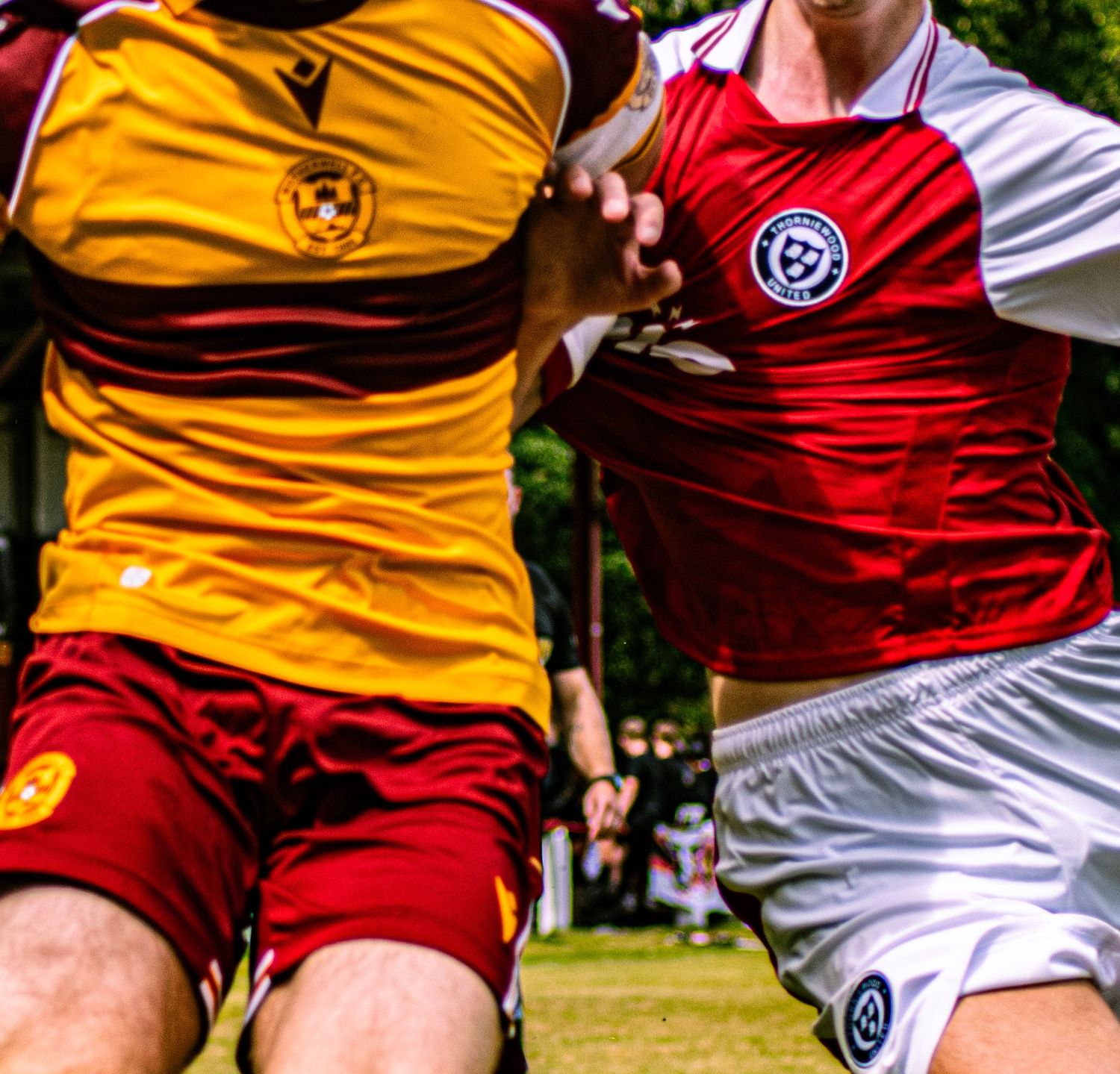 Two soccer players compete for the ball, one in yellow and red kit, the other in red and white striped jersey.
