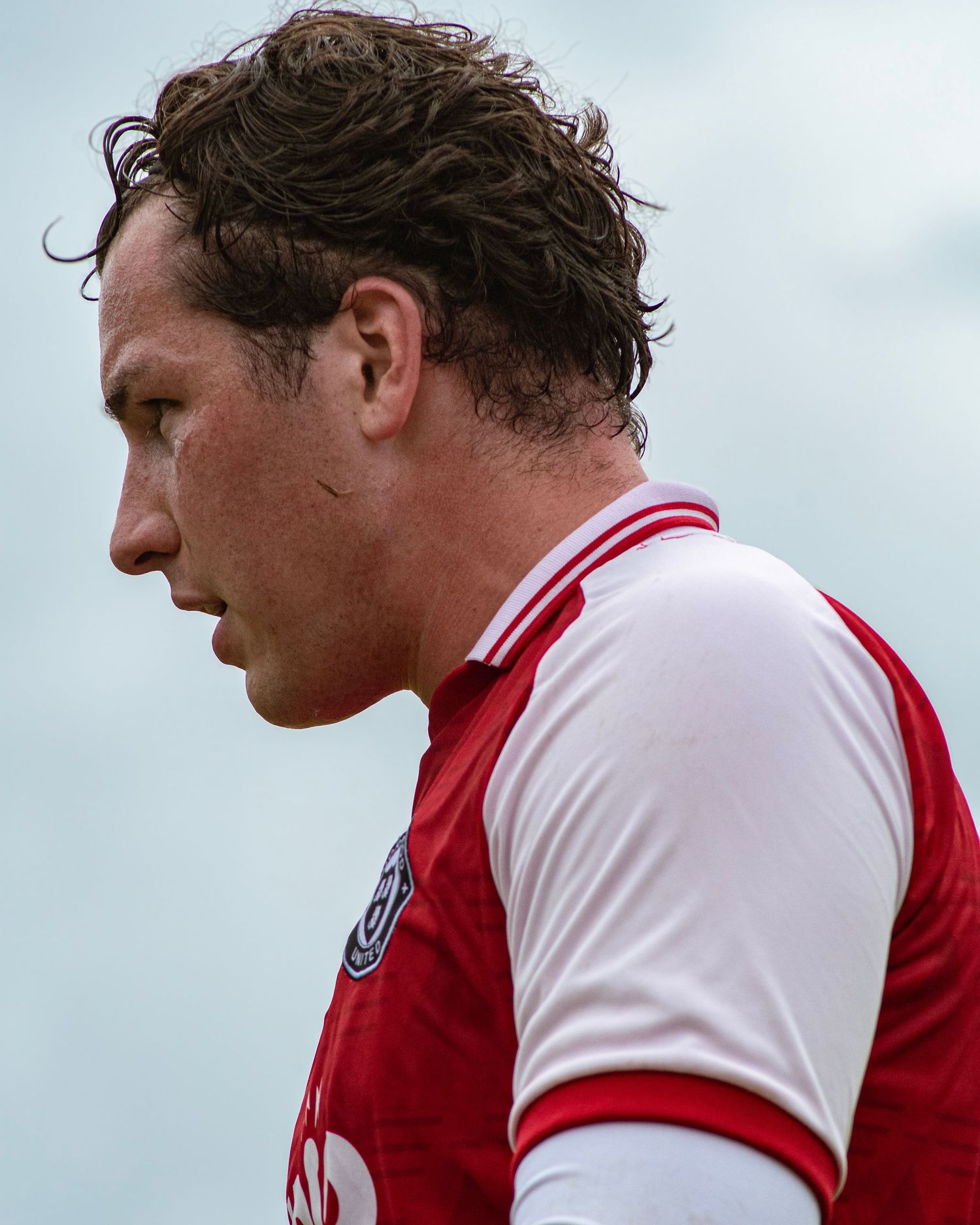 Side profile of a male footballer in a red and white jersey during a game outdoors.