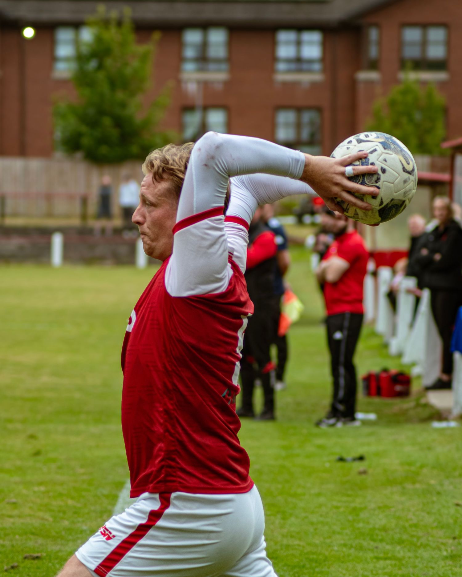 A footballer in red and white kit takes a throw-in during a grassroots football match on a green pitch.