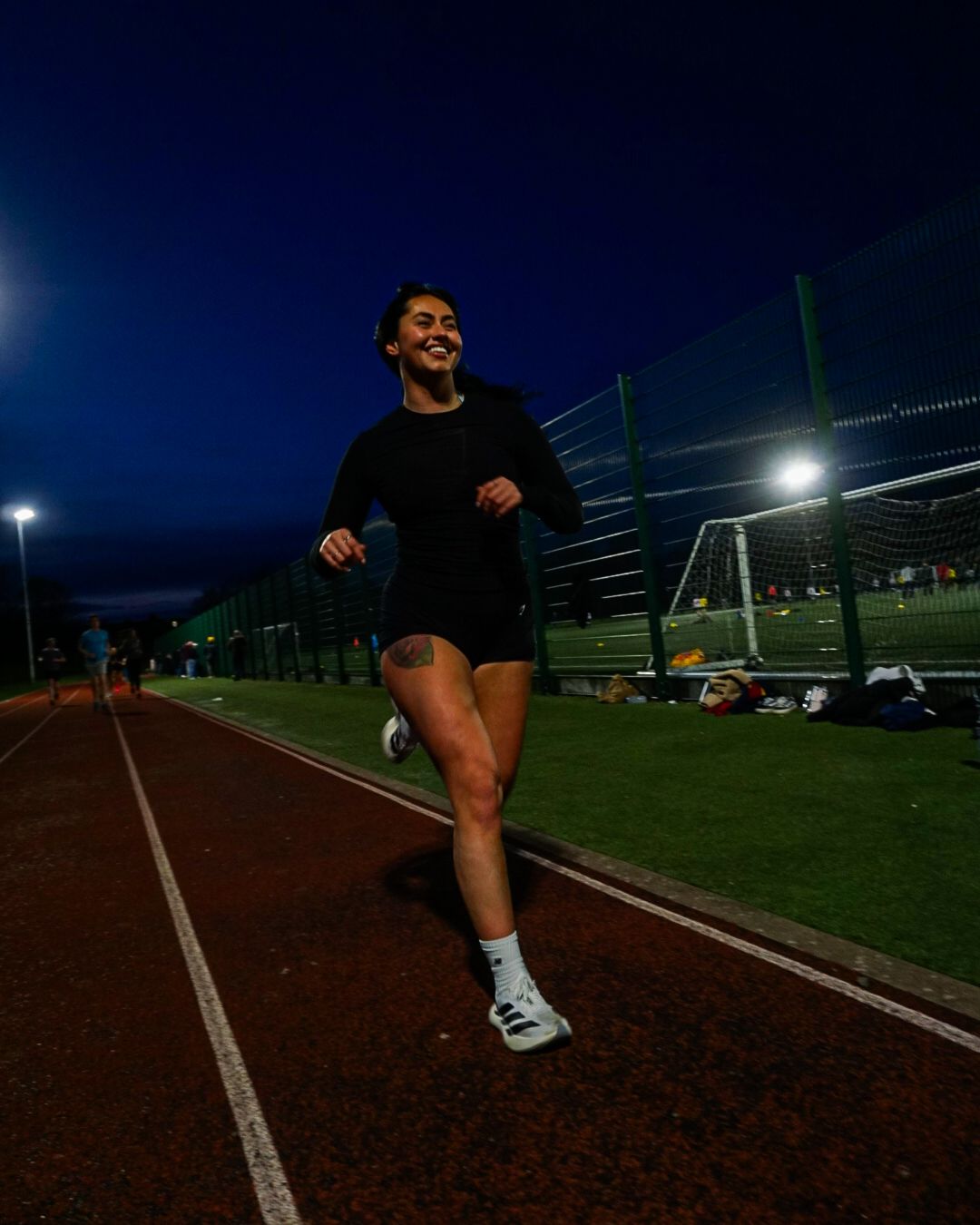 Smiling woman in black athletic wear runs on a floodlit track at night.