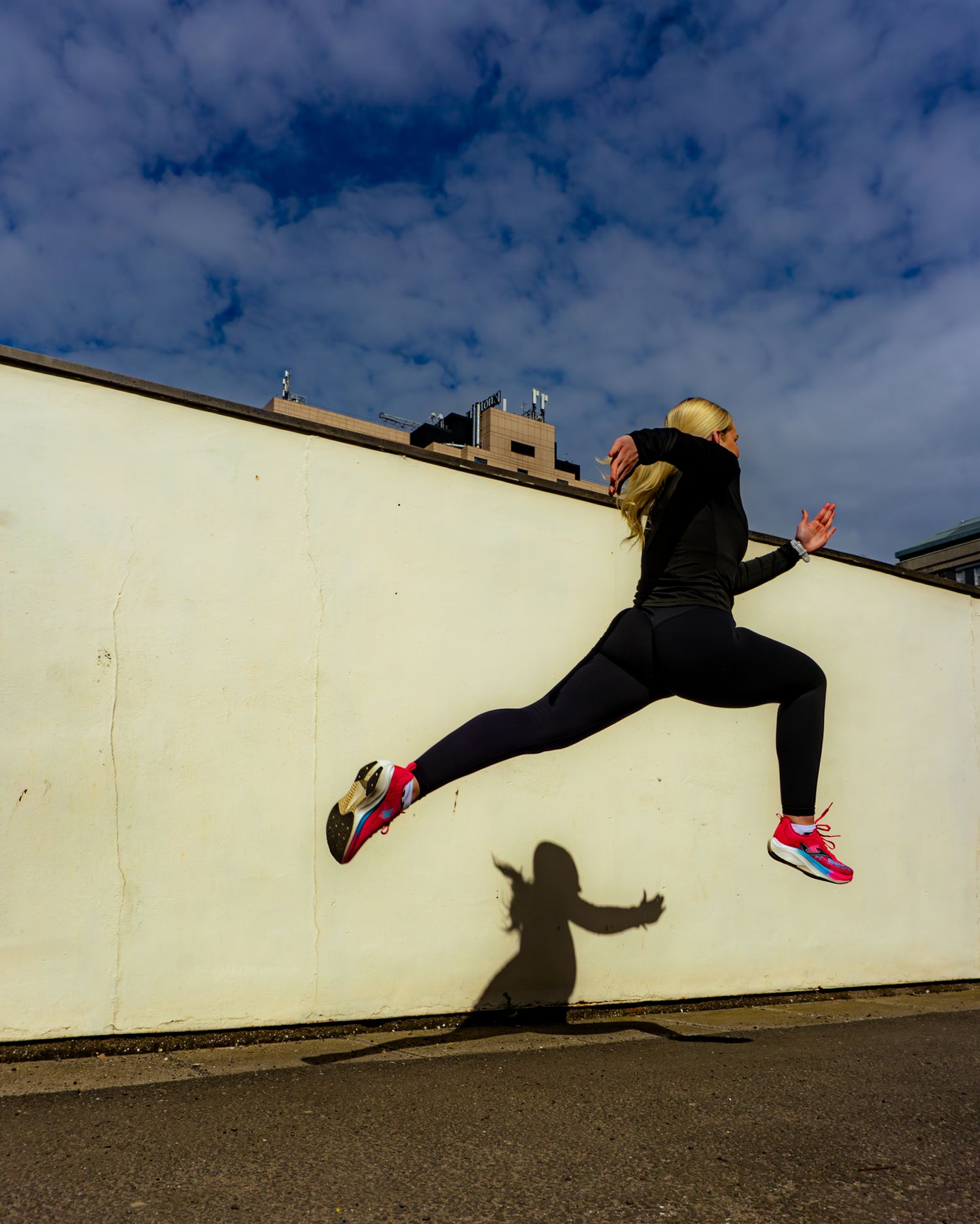 Female athlete in black outfit jumping mid-air against white wall wearing bright pink running shoes.