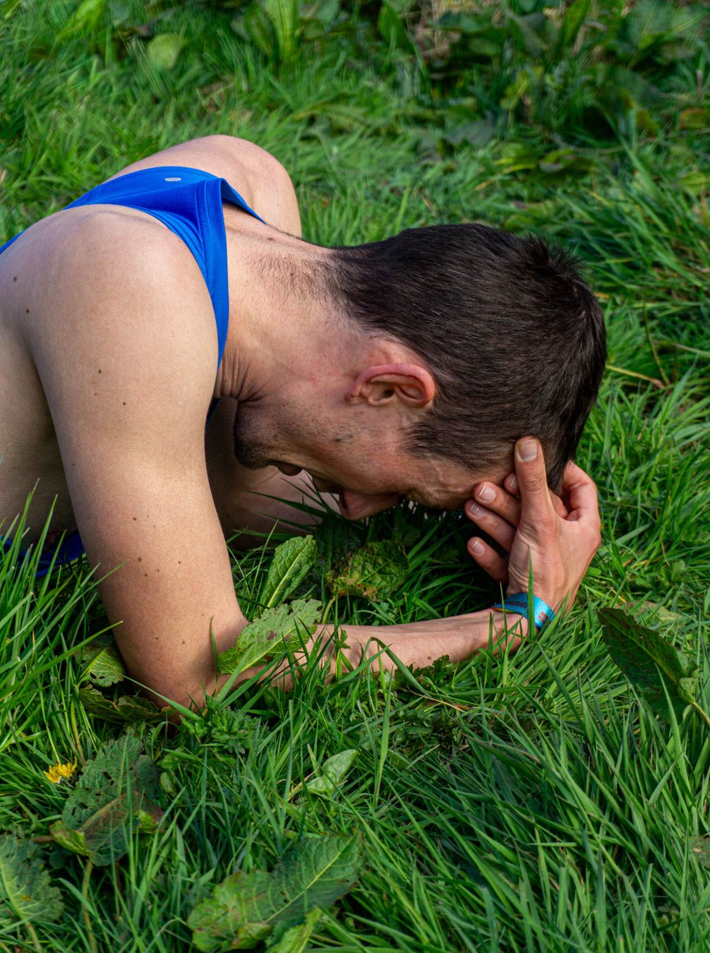 Exhausted male runner in blue vest collapses face down on green grass after a race.