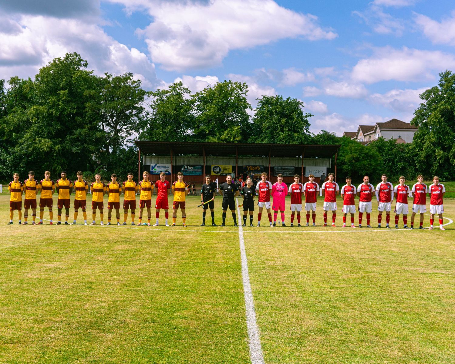 Two youth football teams line up facing each other on sunny grass pitch before kickoff.