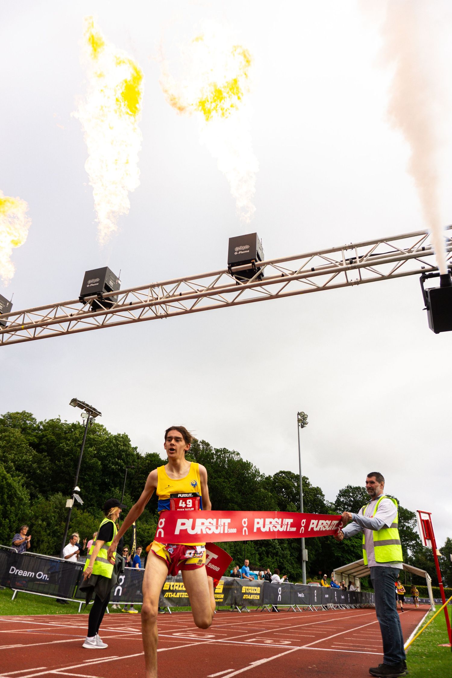 Female athlete crosses finish line at Pursuit race event with dramatic fire cannons blazing overhead on outdoor track.