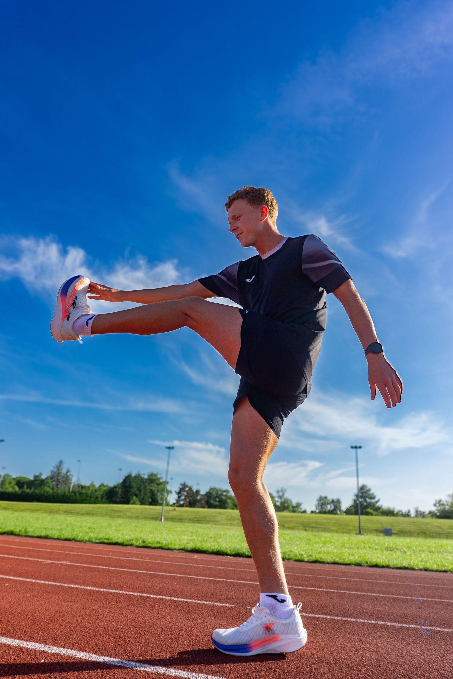Male athlete stretching on a red running track under a bright blue sky, wearing black shorts and white running shoes.