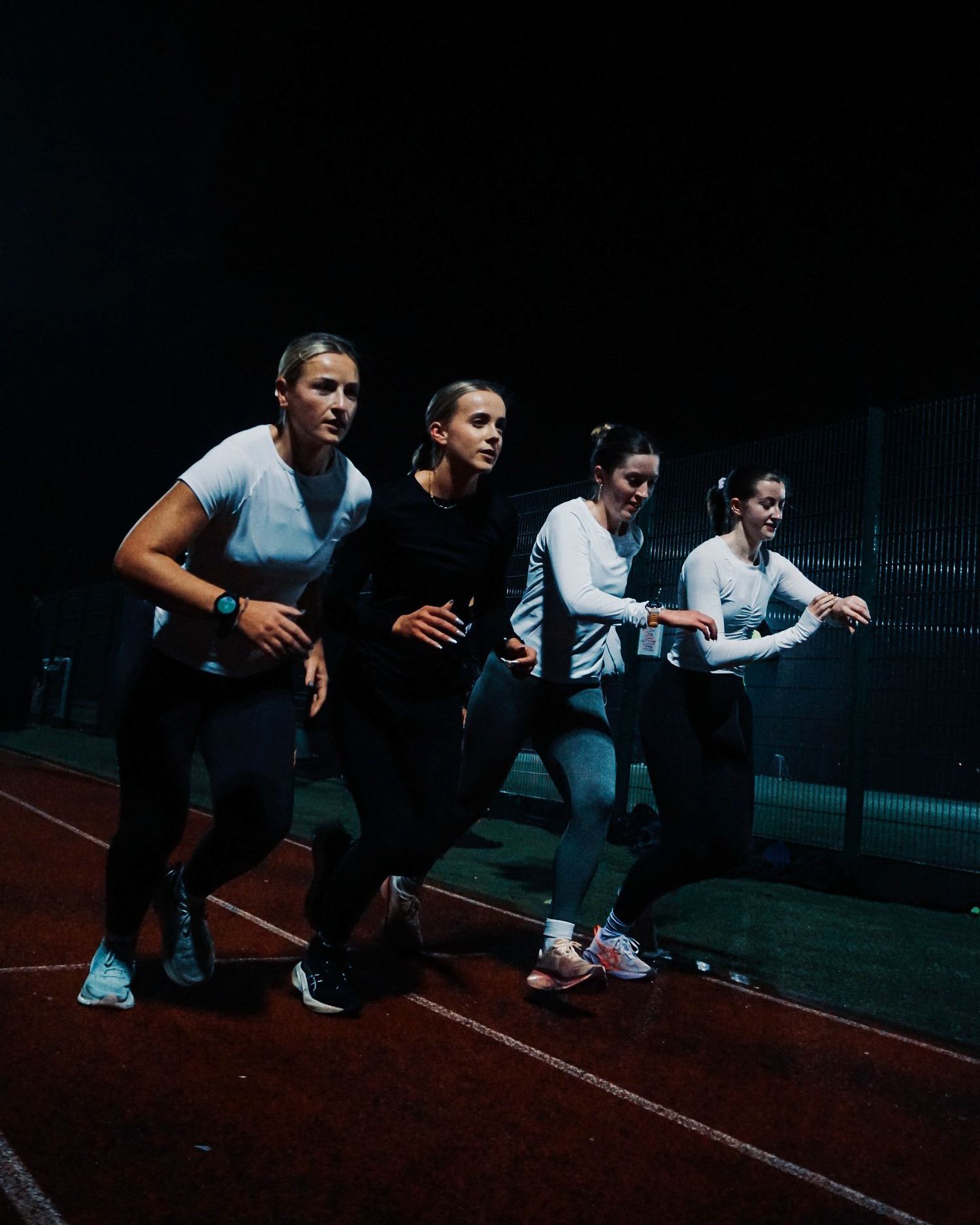 Four female athletes in athletic wear preparing to sprint on an indoor running track at night.